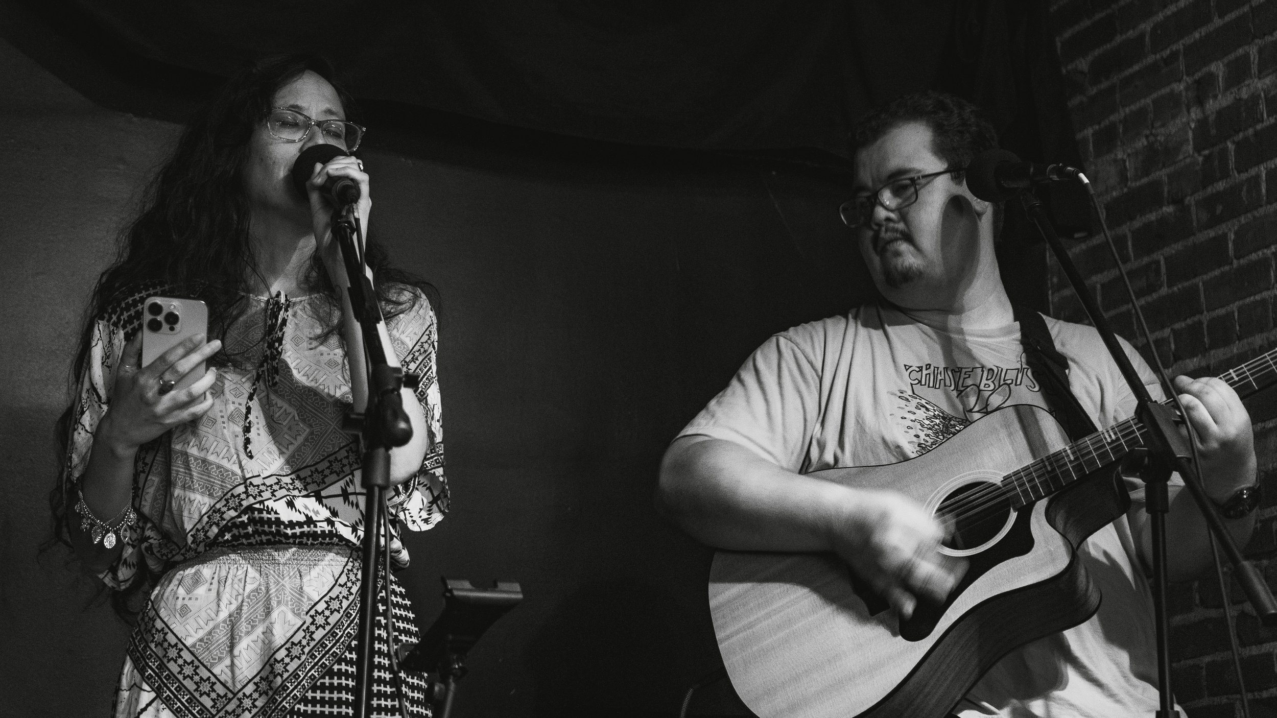 A woman singing into a microphone while holding a smartphone, and a man playing an acoustic guitar on a stage with a brick wall background.