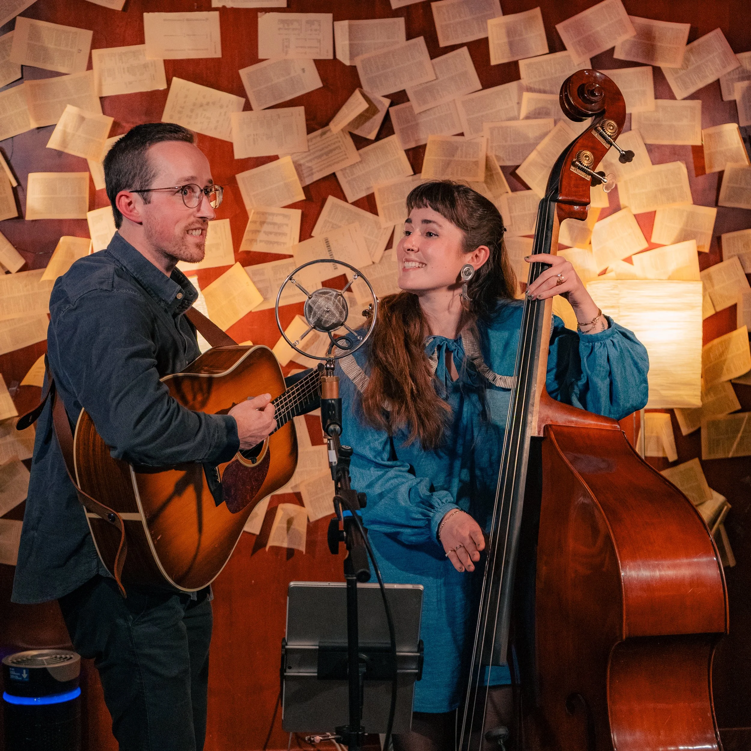 A man playing an acoustic guitar and singing into a vintage-style microphone, and a woman playing a double bass and smiling, performing in a cozy room with walls decorated with open books.