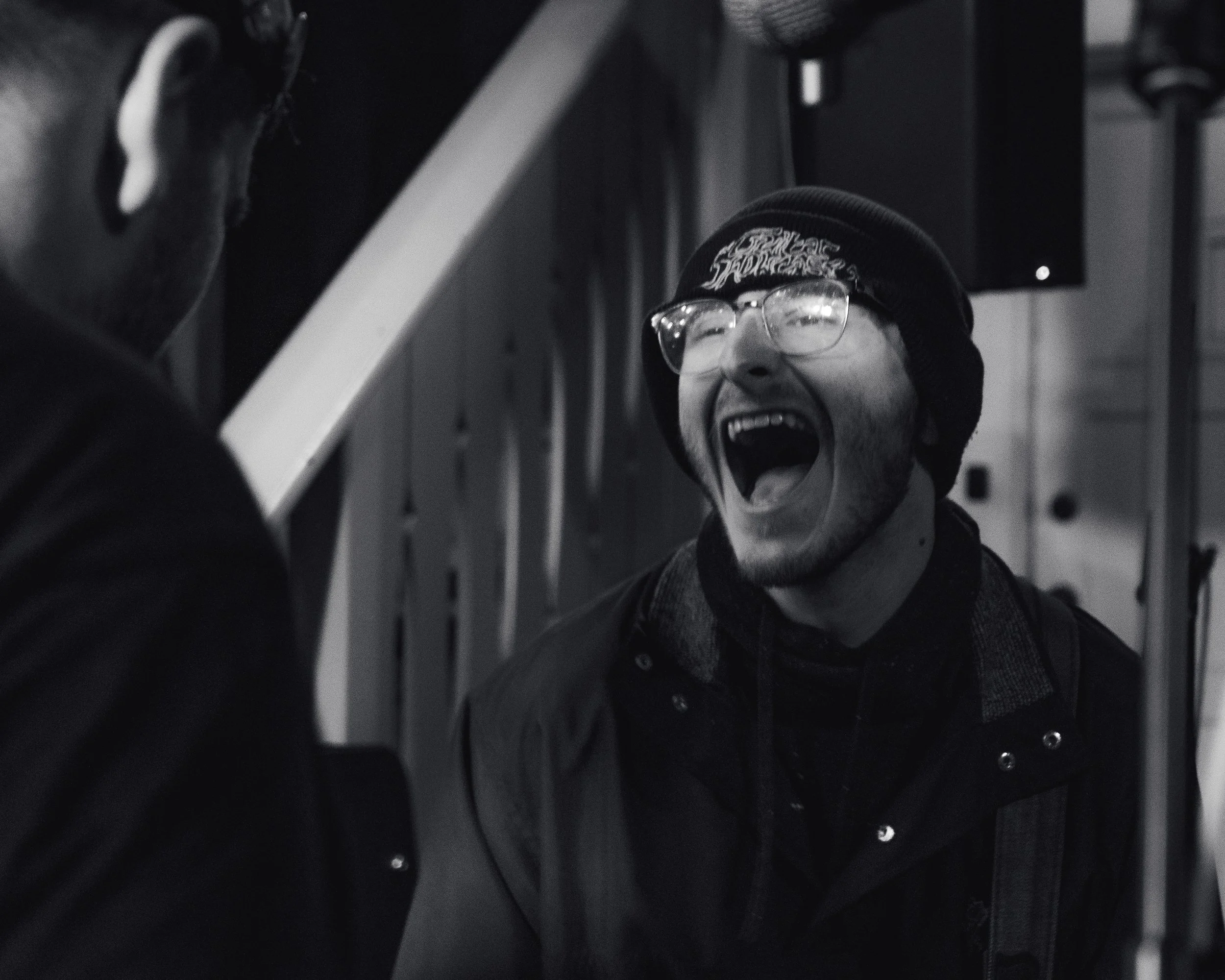 A man is smiling and laughing, wearing glasses, a beanie, and a jacket, in a black and white photo, with a background of a wood interior.