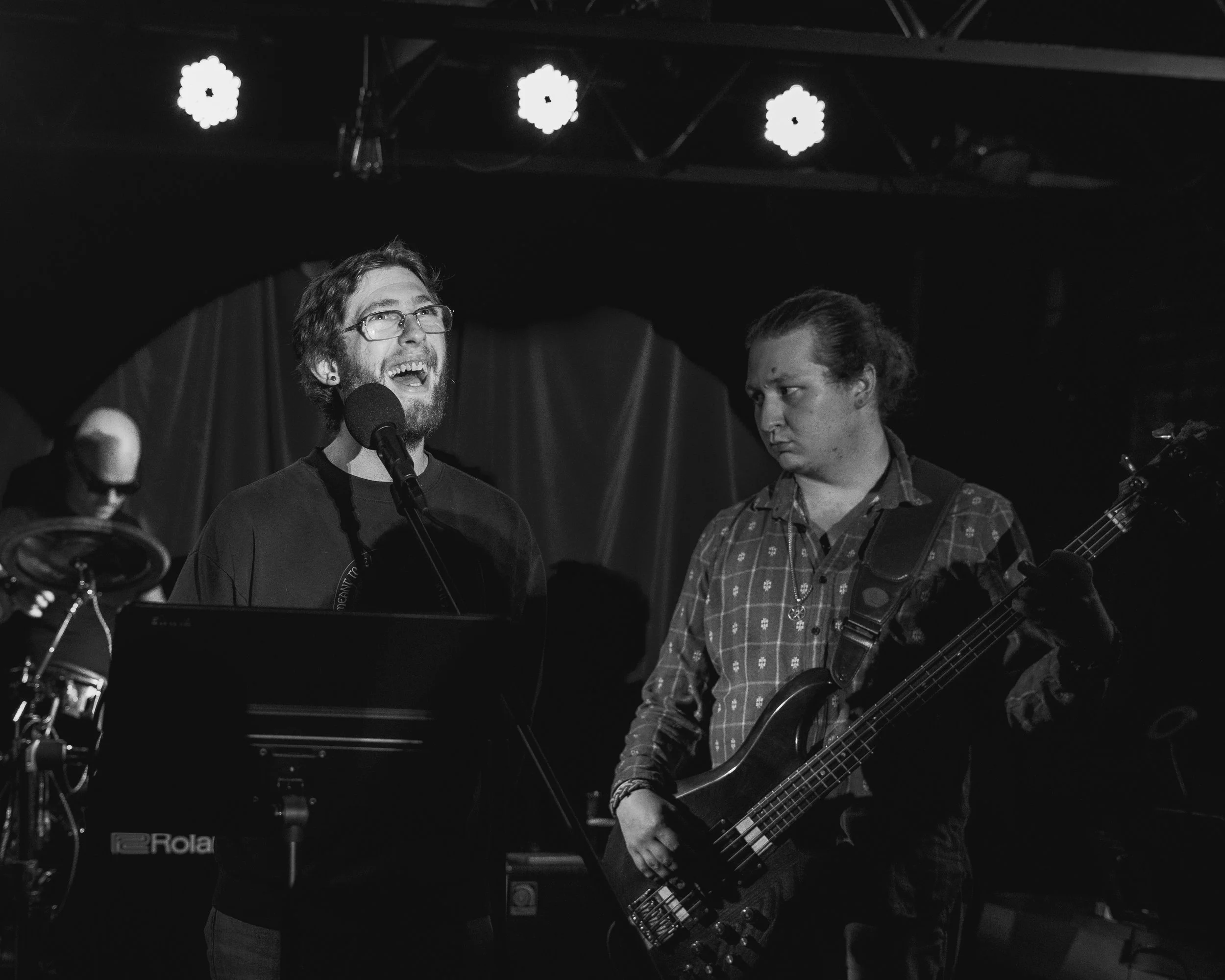 Two musicians on stage with a man singing into a microphone and a bassist playing. A drummer is in the background. The stage has spotlights and draped fabric.