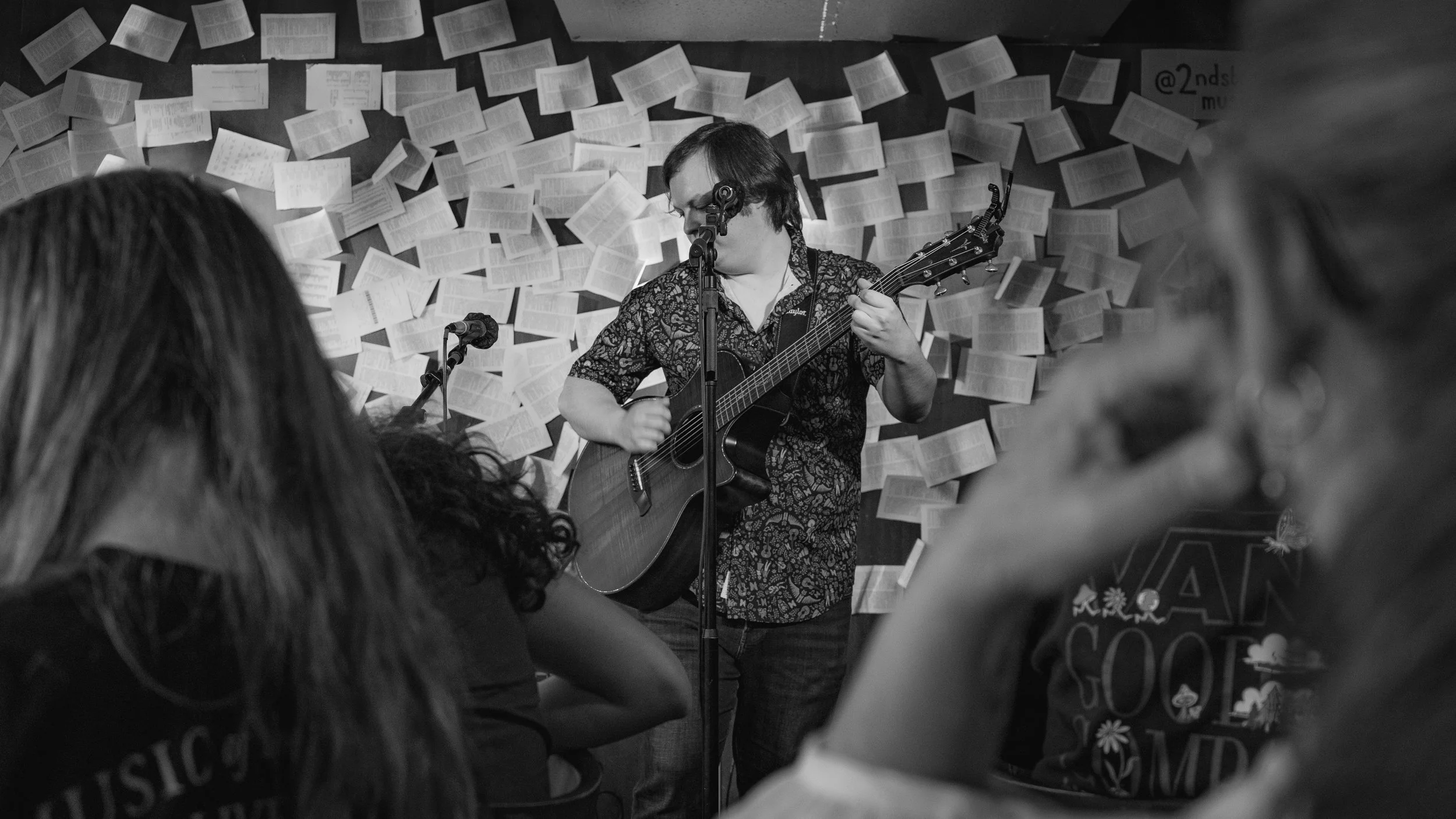 A musician with glasses and wearing a patterned shirt performs with an acoustic guitar and sings into a microphone on a small stage with a backdrop of open books on a wall and some audience members in the foreground.