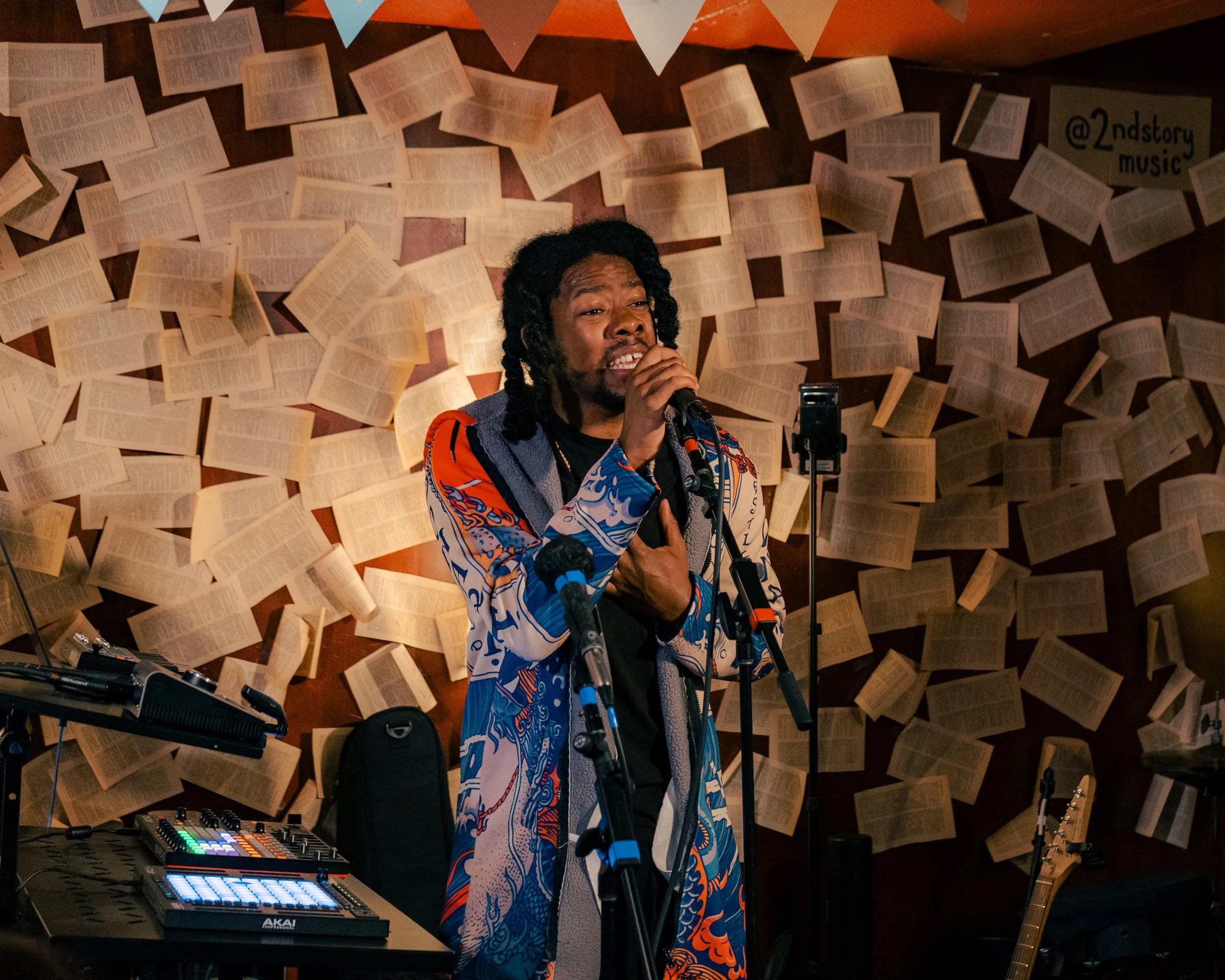 A man singing into a microphone in a room decorated with pages from books and banners on the wall.