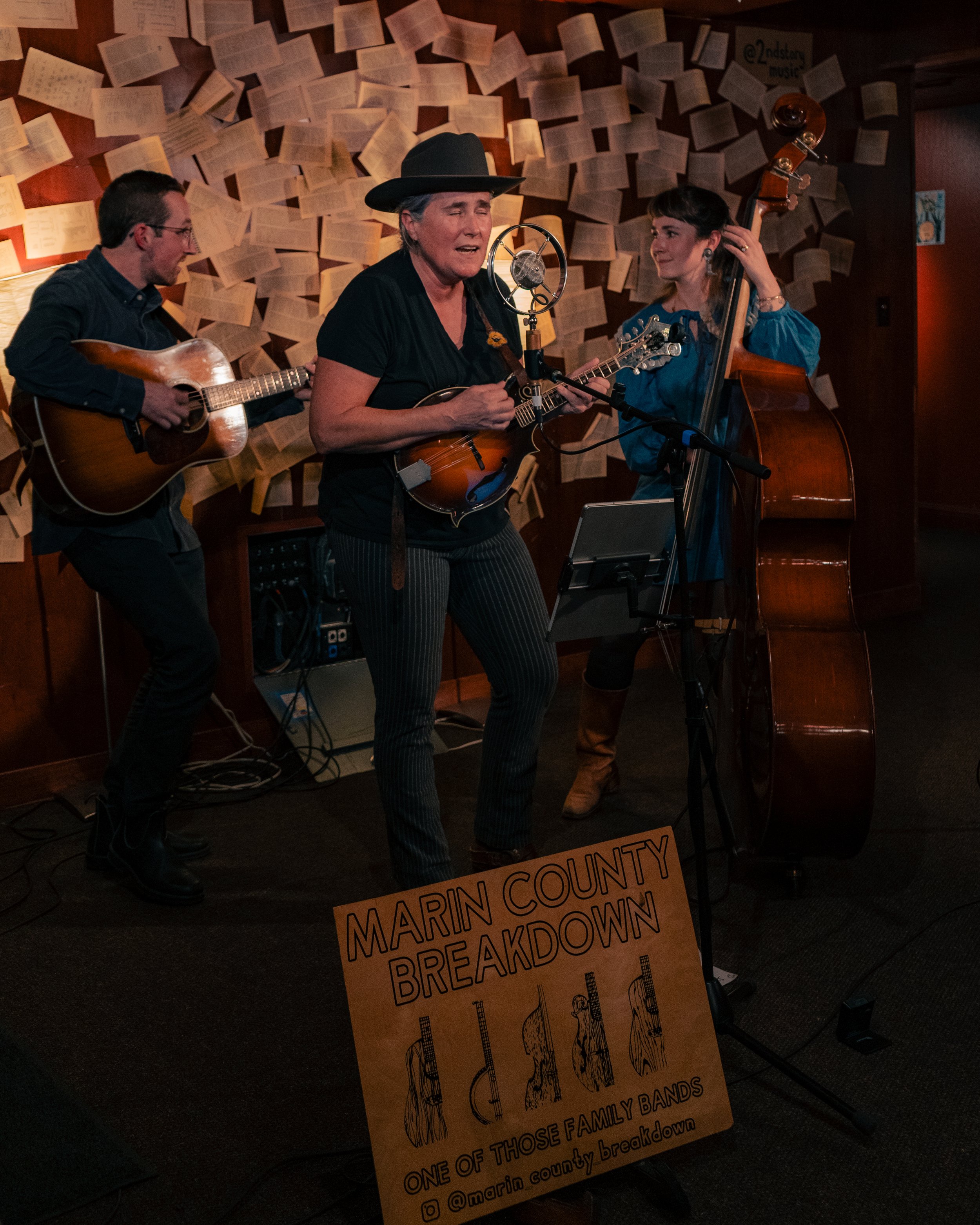 Three musicians performing on stage, two women and one man, with a backdrop of numerous open books on the wall, and a sign reading "Marin County Breakdown" in front of them.
