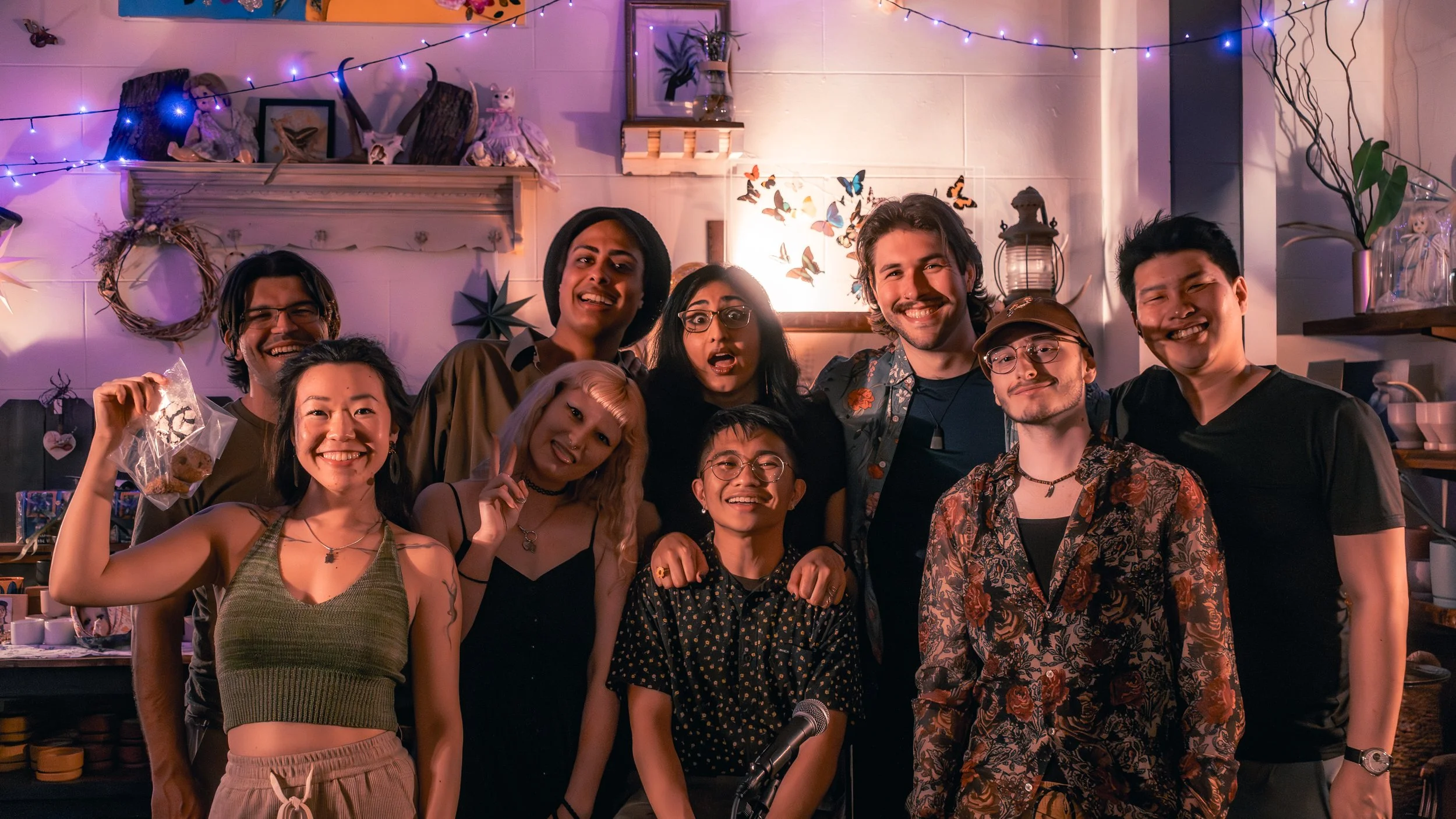 A group of eleven diverse friends smiling and posing for a photo indoors, with colorful fairy lights and decorations in the background.
