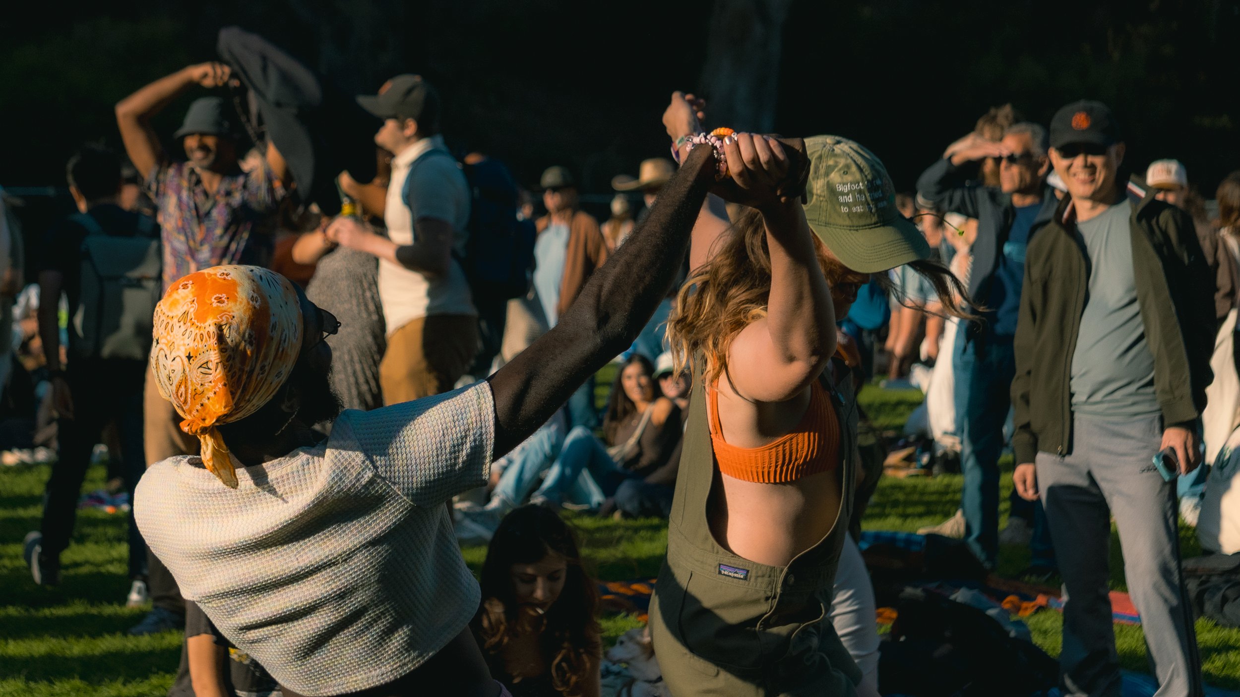 Two women dancing at an outdoor event with a crowd in the background on a grassy field.