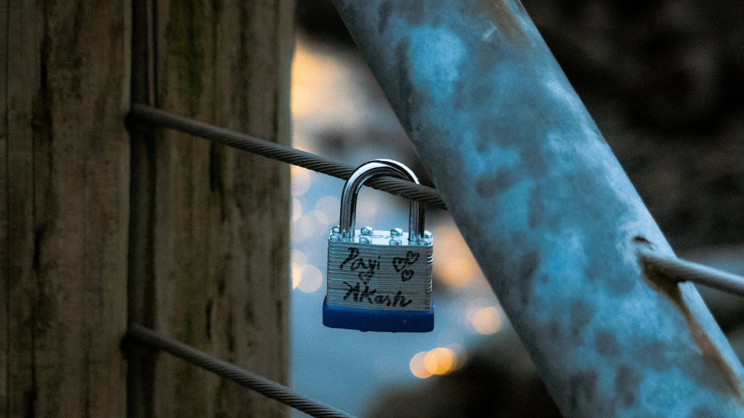 A padlock attached to a metal cable on a bridge or railing, with handwritten names and hearts, at sunset or dusk.