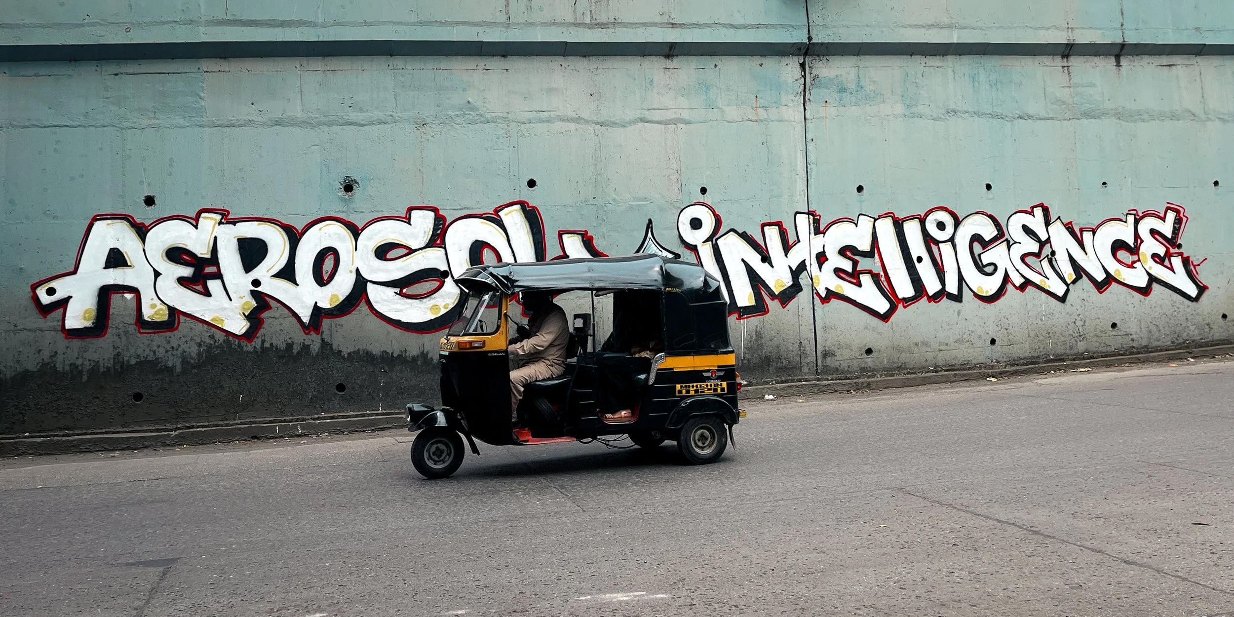 Graffiti art on a wall with the words 'AEROSOL INTELLIGENCE' painted in bold, colorful letters. A black and yellow auto rickshaw with a driver seated inside is passing by in front of the wall.