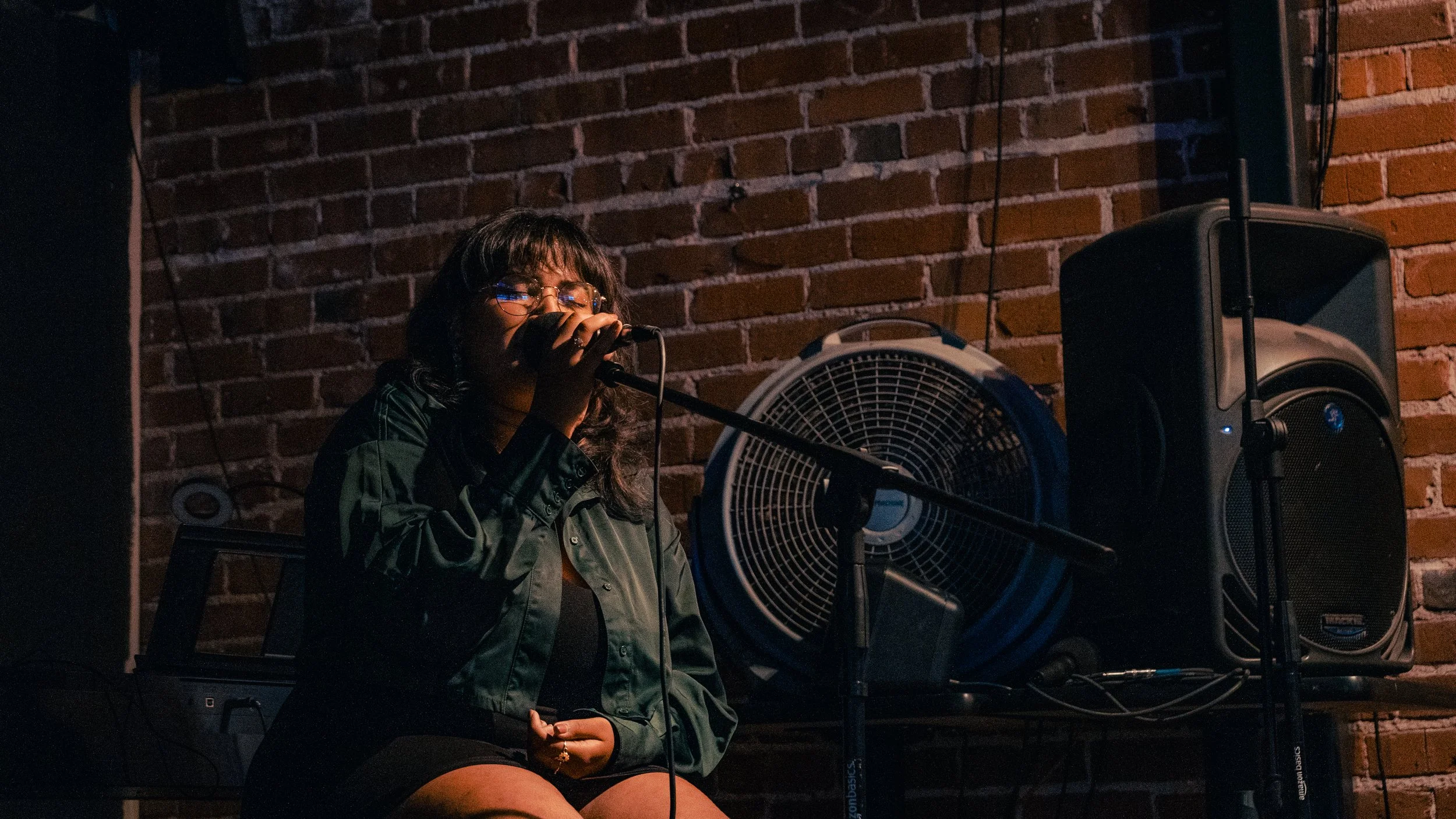 A woman with glasses and dark hair is singing or speaking into a microphone at an indoor venue, with a brick wall in the background, a large fan, and speakers nearby.