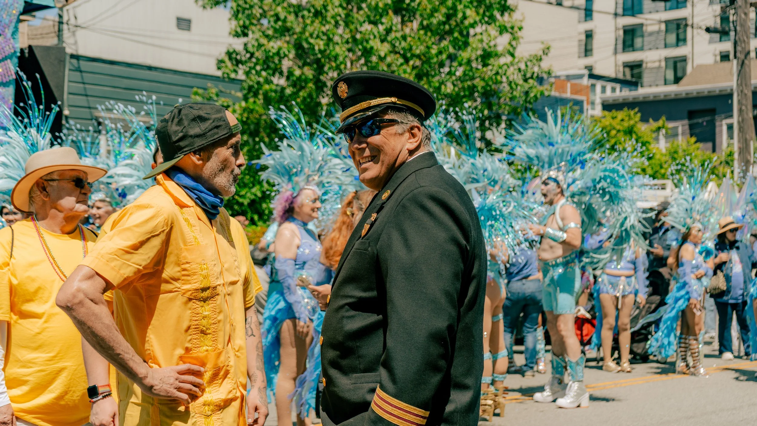 A man wearing a pilot uniform smiling and talking outdoors during a parade with people dressed in blue costumes with feathered headdresses in the background.