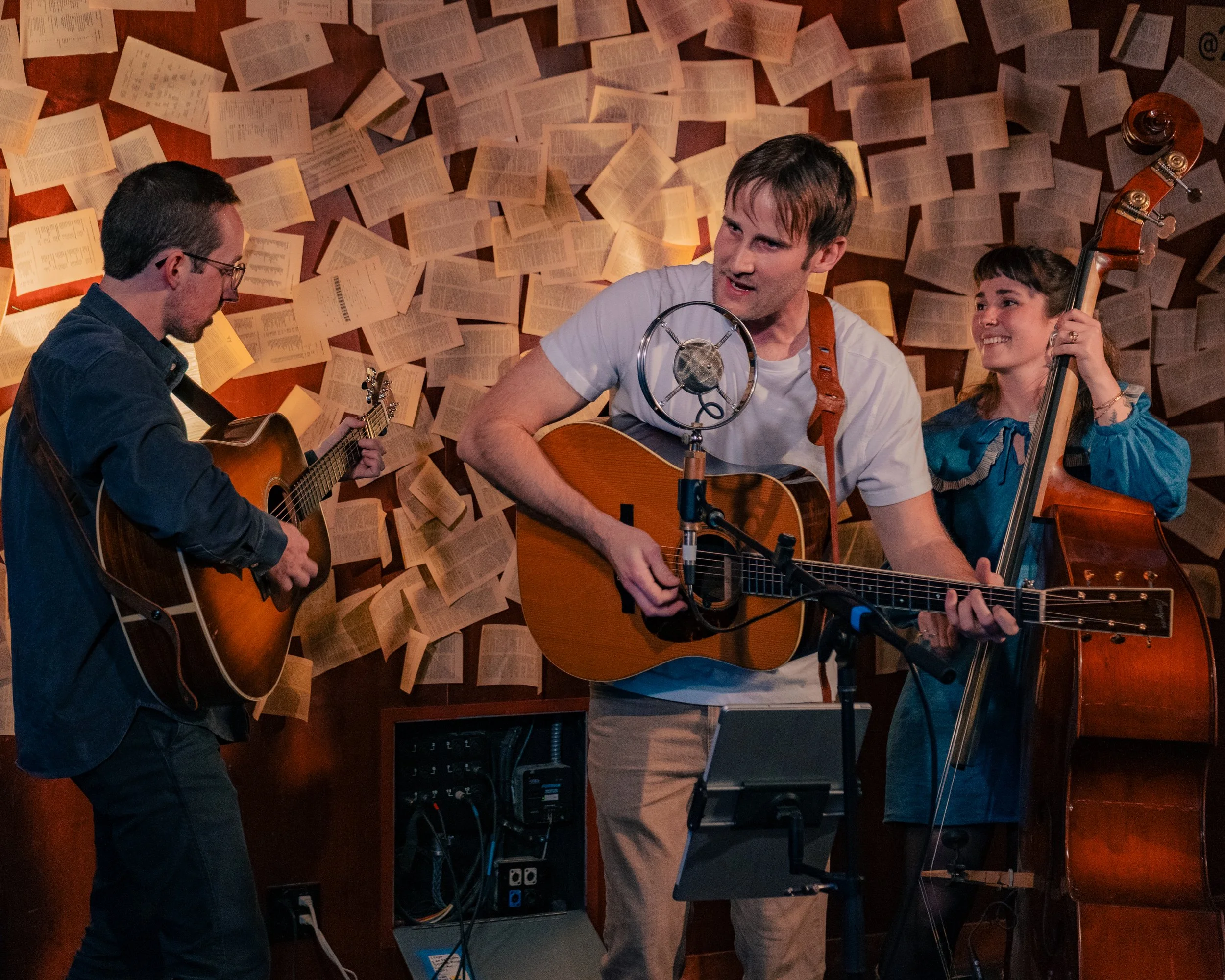 Three people playing guitars and a double bass in a room decorated with pages from books on the walls, with a vintage microphone in front of the man in the middle.