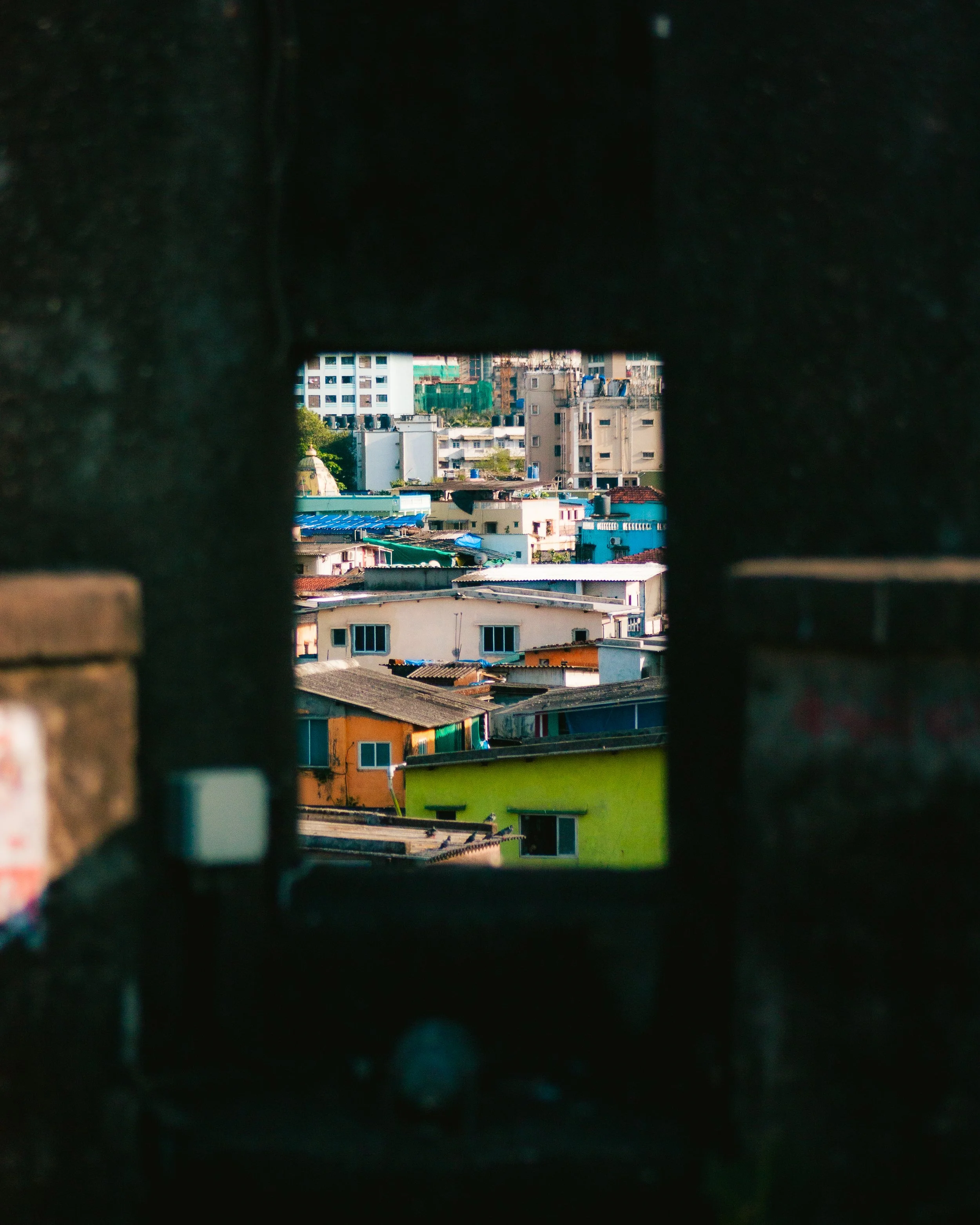 Cityscape viewed through a small opening in a brick wall, showing colorful buildings and taller structures in the background.