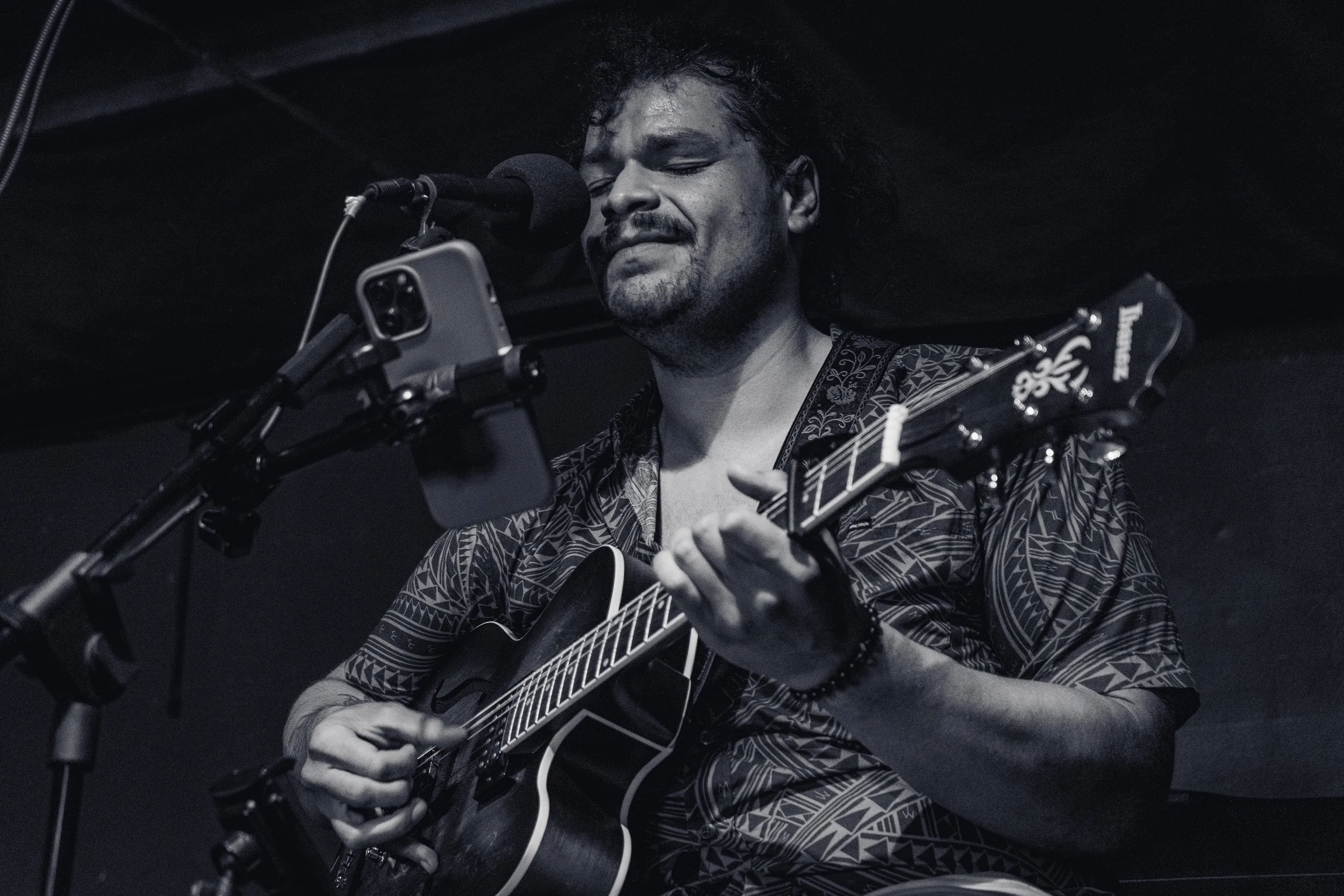 A male musician singing into a microphone while playing an acoustic guitar, with a phone mounted on a stand recording him, in a dimly lit setting.