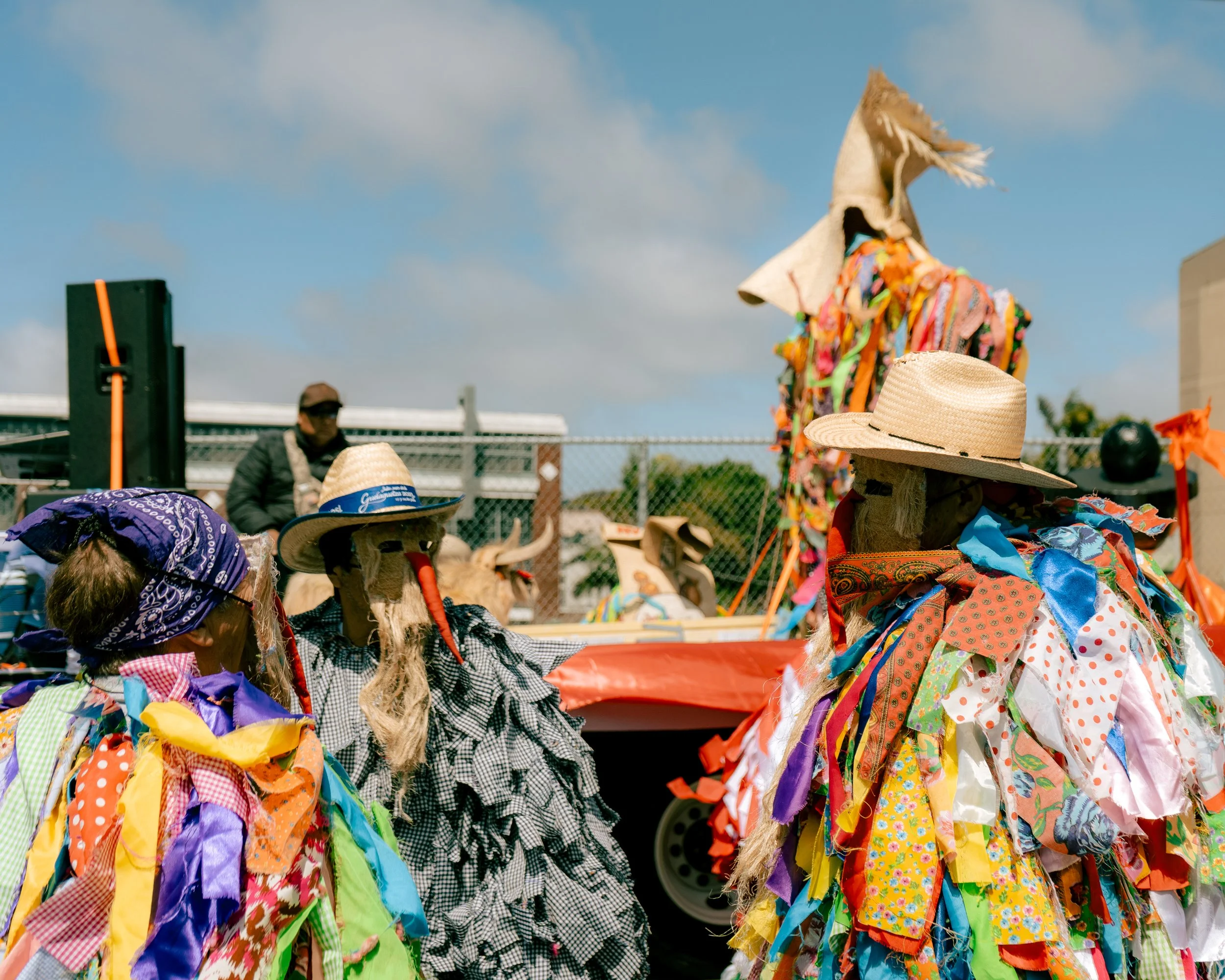 Colorful traditional costumes with fabric strips and straw hats worn by people in fabric dolls costumes in front of a stage with a person speaking, a fence, and a large puppet or figure dressed in colorful fabric in the background on a sunny day.
