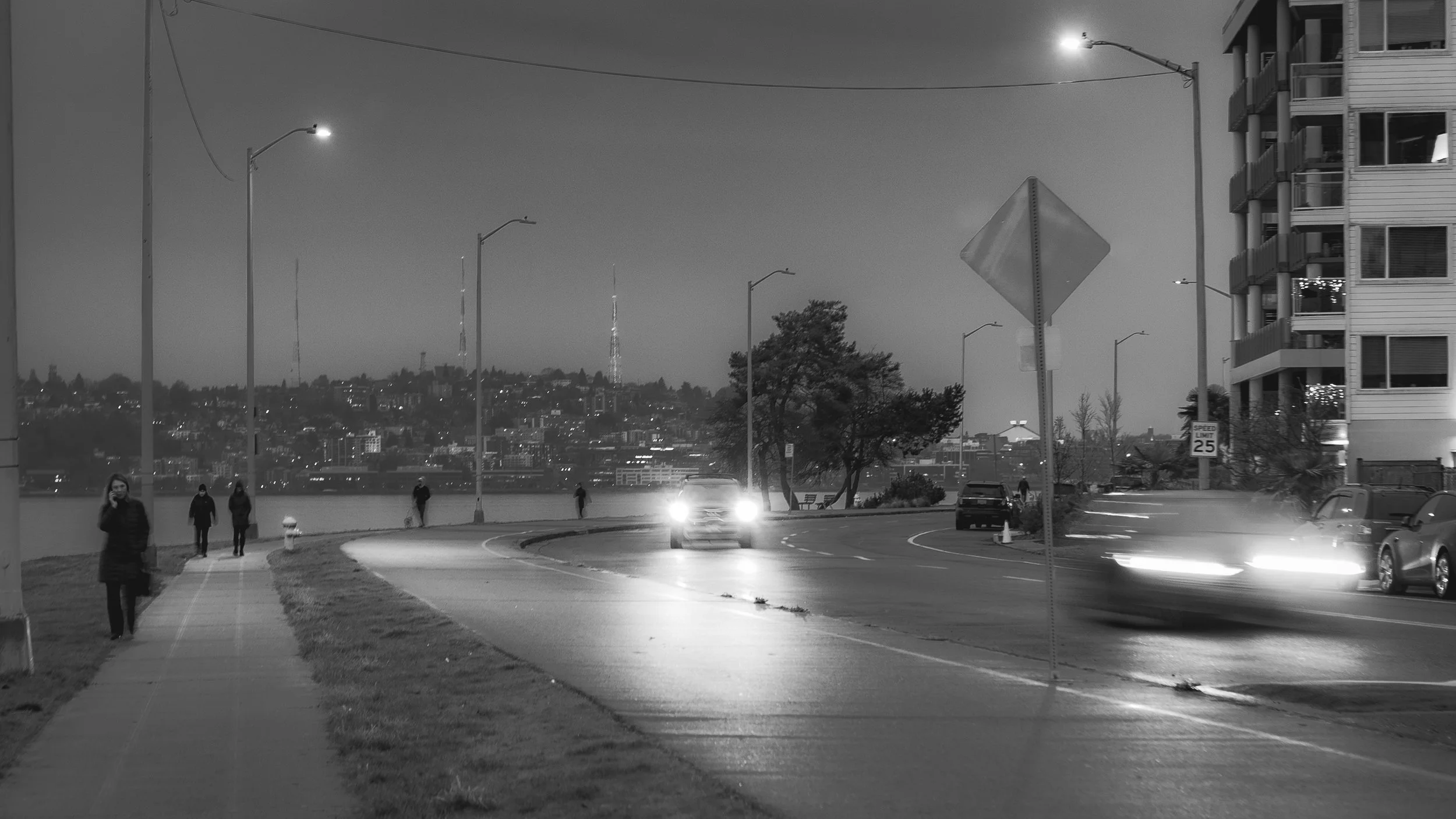 A black and white photo of a city street at night with streetlights, a few pedestrians walking on the sidewalk, and cars on the road, some with headlights on and in motion.