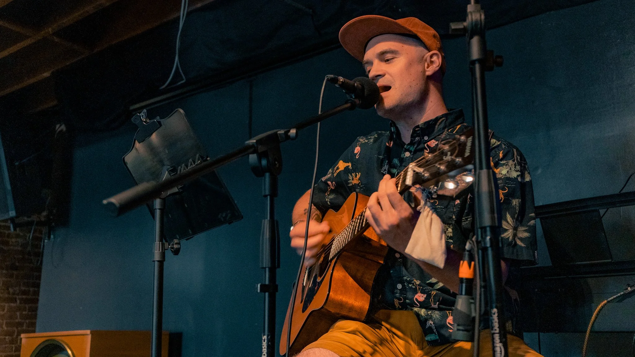 A man playing an acoustic guitar and singing into a microphone on stage, wearing a patterned shirt and a hat, with audio equipment and lighting in the background.