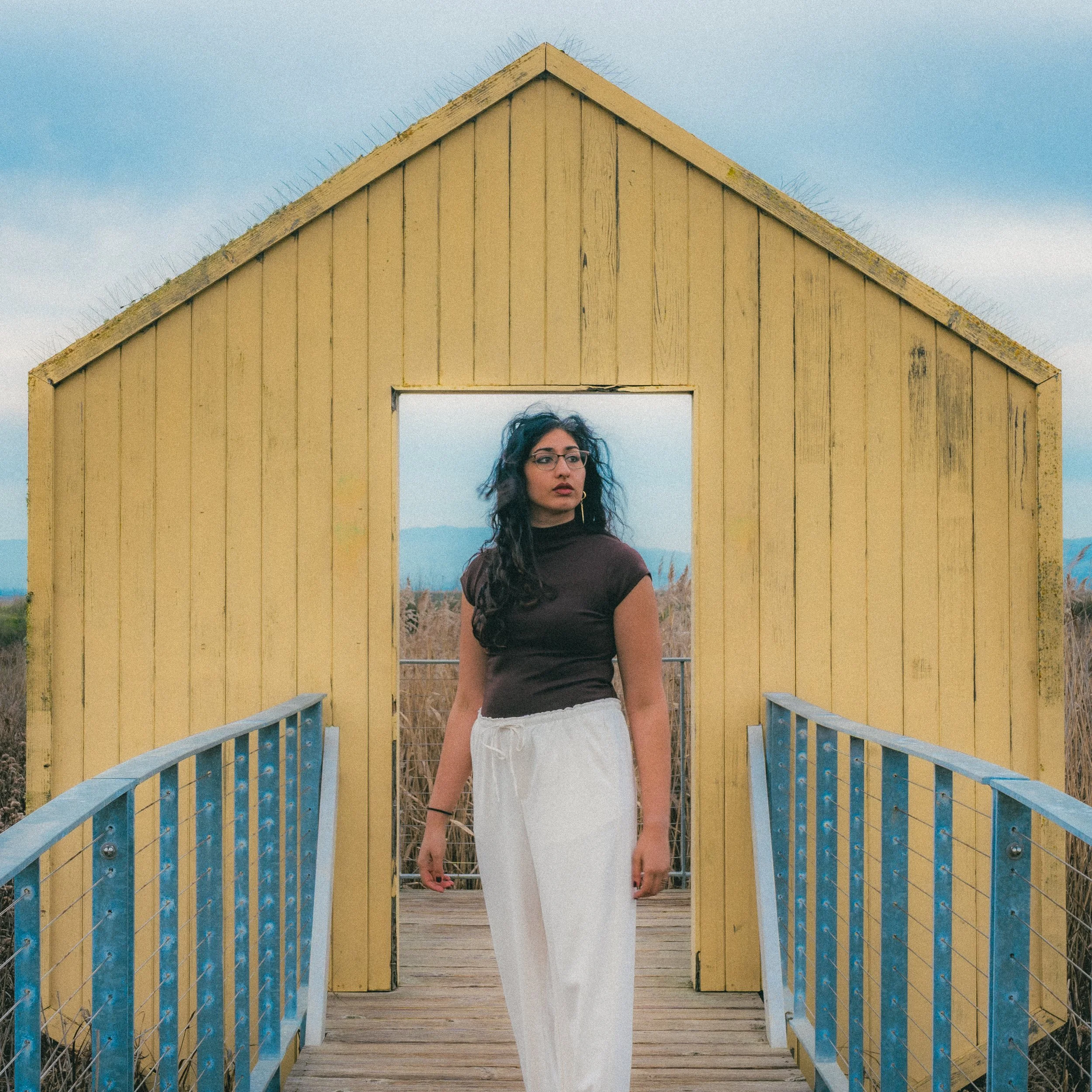 A woman with curly dark hair, glasses, and earrings stands on a wooden pathway in front of a yellow wooden structure resembling a small house with a steep roof, with blue sky and mountains in the background.