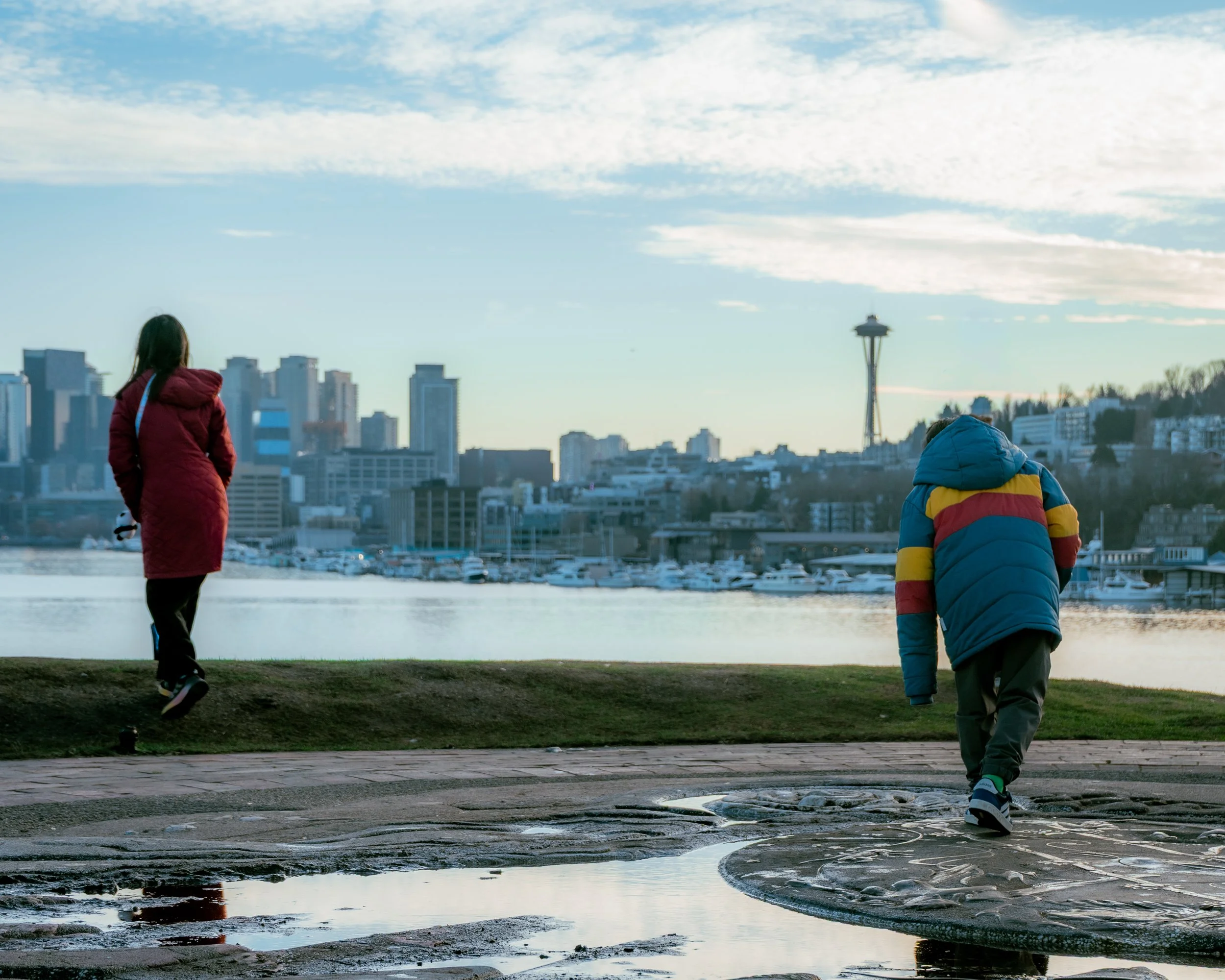 A woman in a red coat walking near the water on the left, and a boy in a colorful winter jacket on the right, over puddles on a paved surface, with the Seattle skyline and the Space Needle in the background.