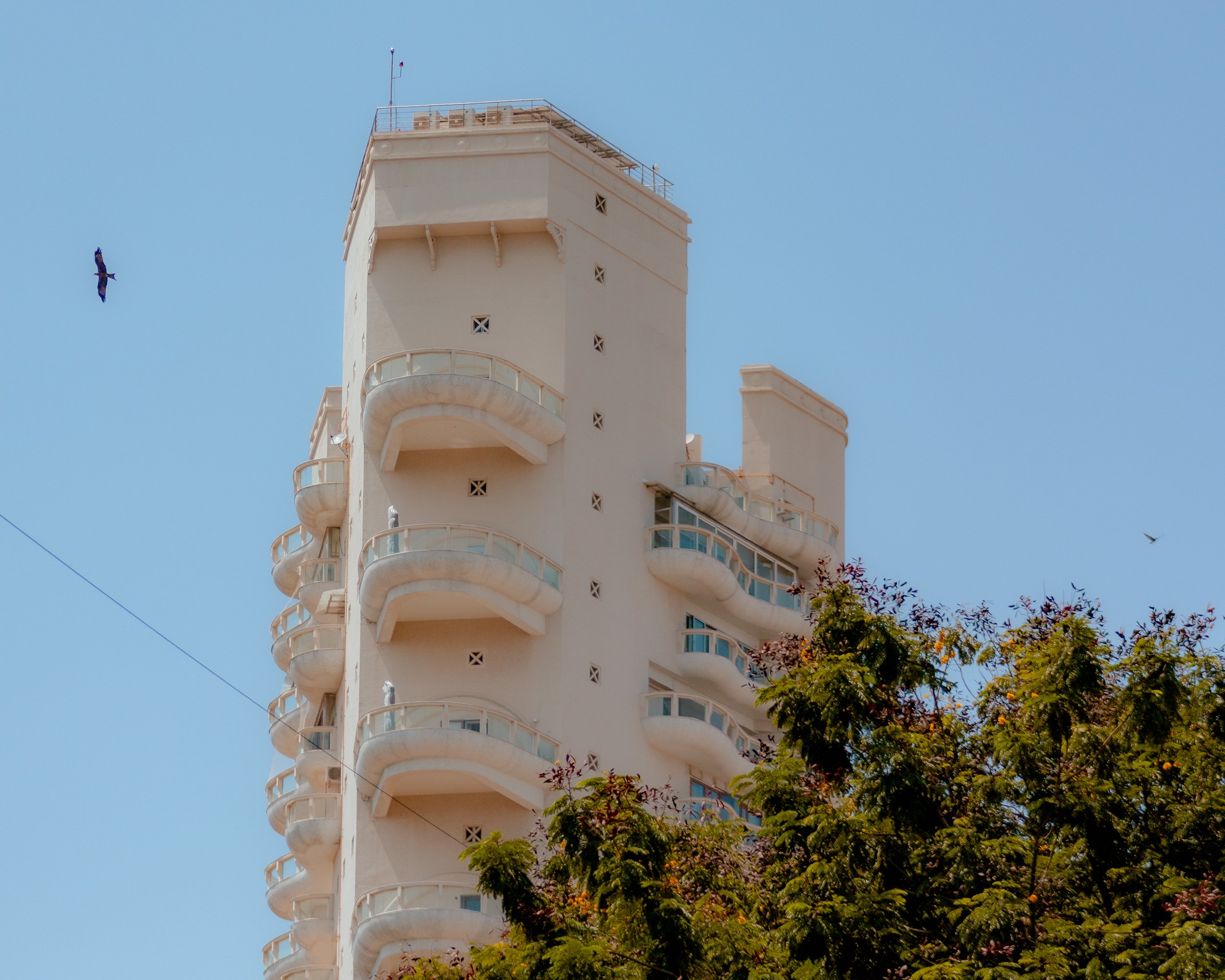 A tall white building with rounded balconies on each floor, partially obscured by trees in the foreground, set against a clear blue sky with a bird flying nearby.