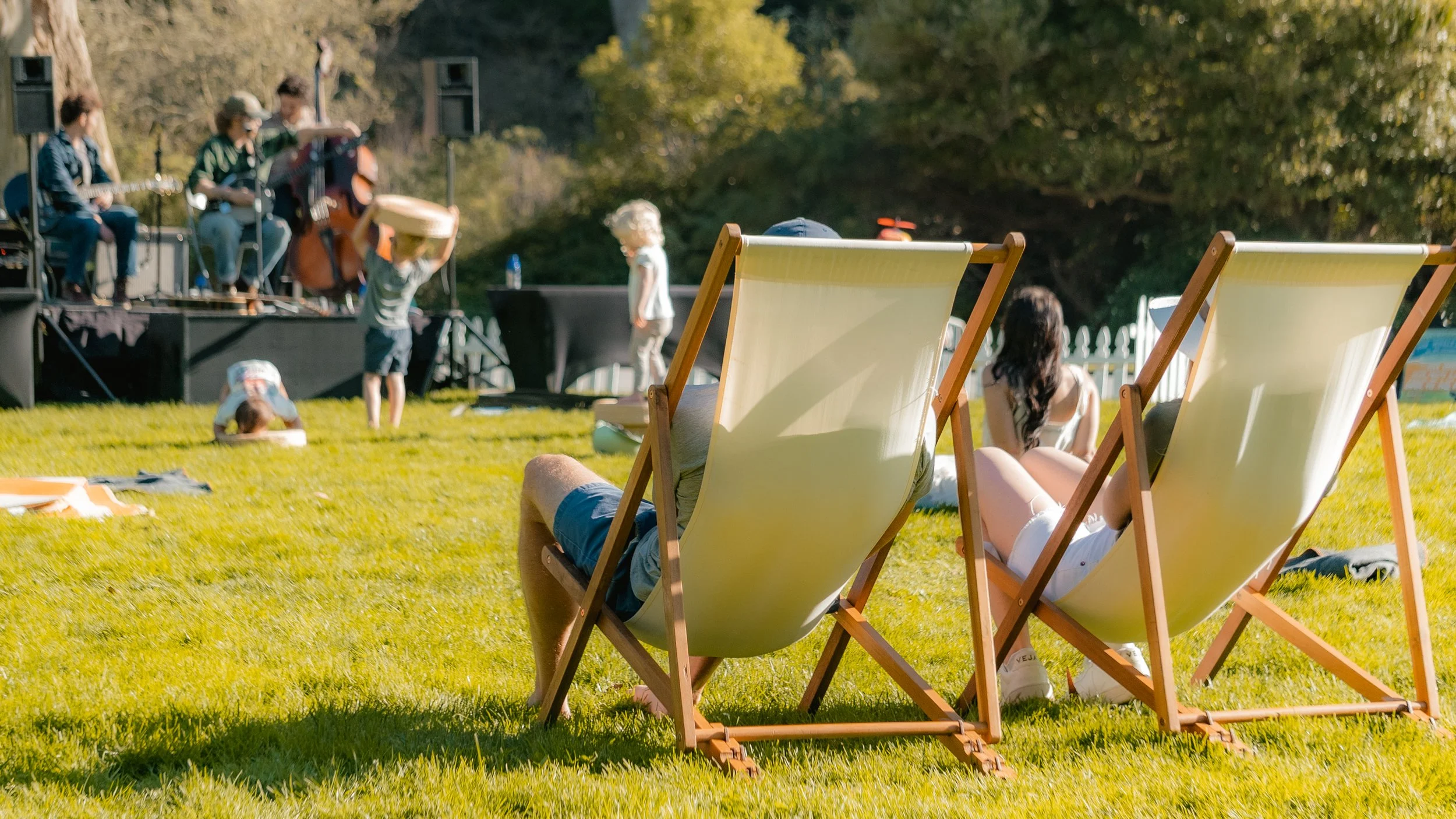 People relaxing on lawn chairs watching a live outdoor musical performance on a stage with musicians and children playing.