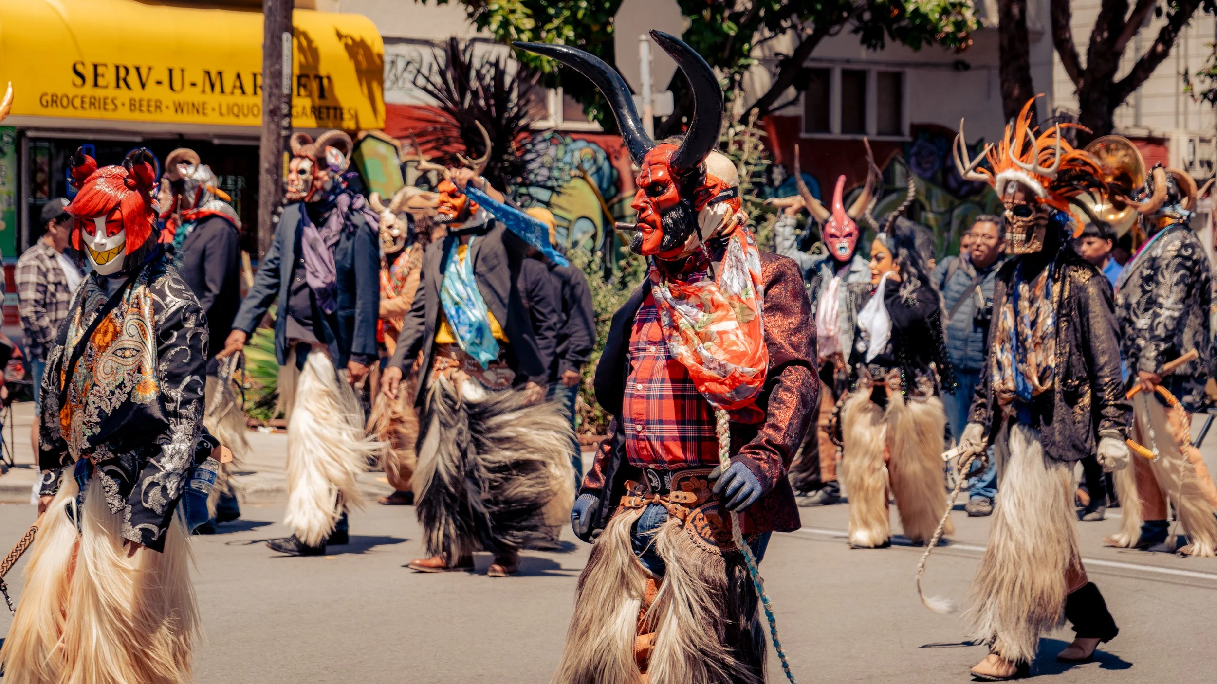 A group of people dressed in elaborate costumes with masks and large furry leg coverings are participating in a parade on a city street, with onlookers and buildings in the background.
