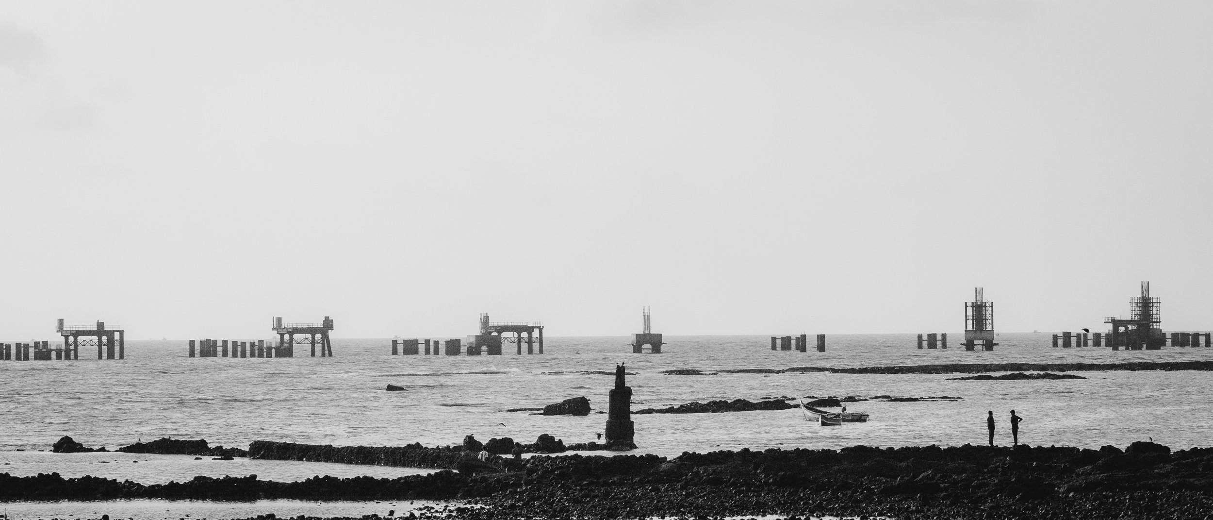 Black and white photo of a seascape showing several construction platforms in the water, with two people standing on the rocky shoreline and a small boat nearby.