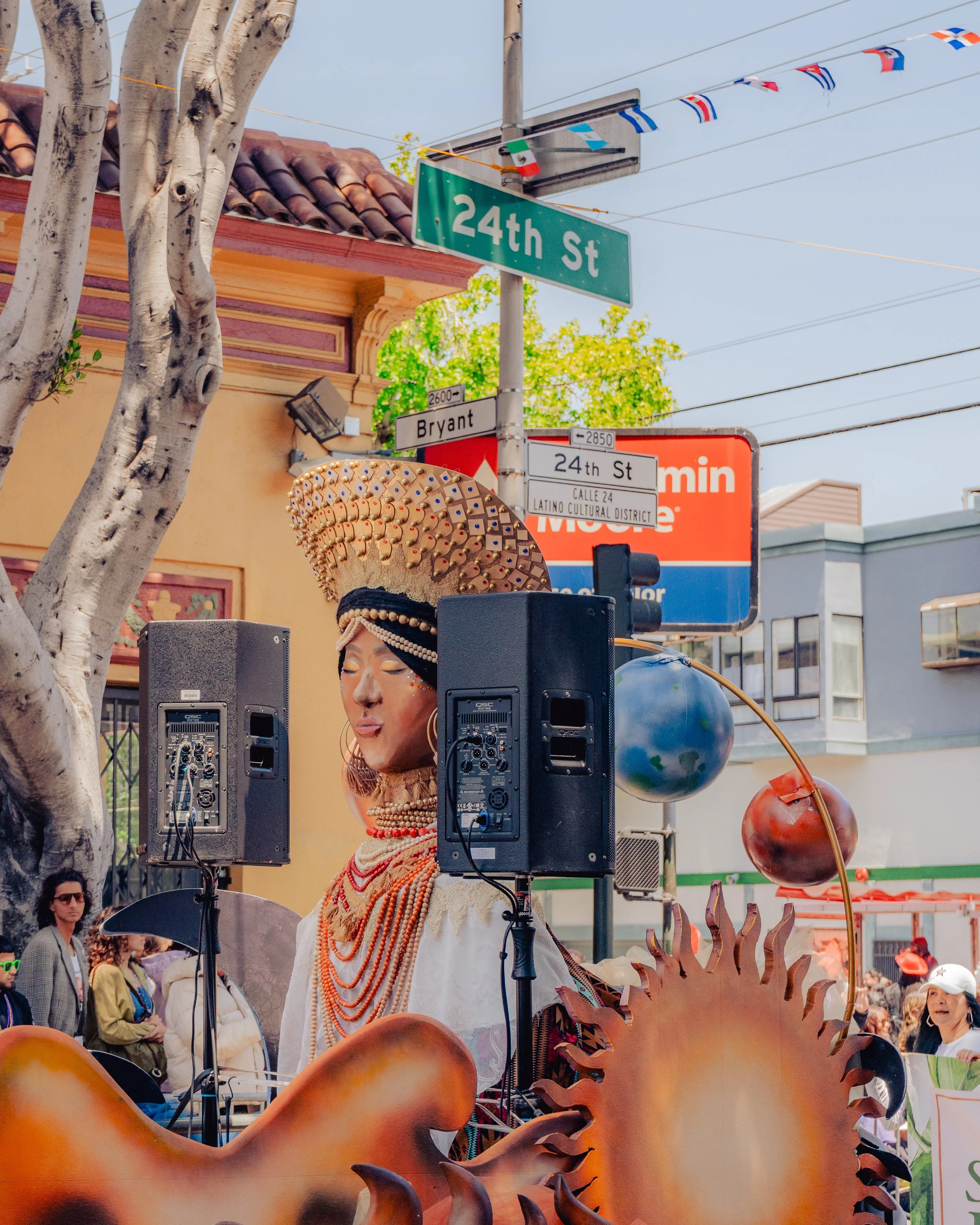 A woman in traditional indigenous attire with a detailed headdress speaking into a microphone at a street event, with street signs indicating 24th Street and Bryant Street visible in the background.