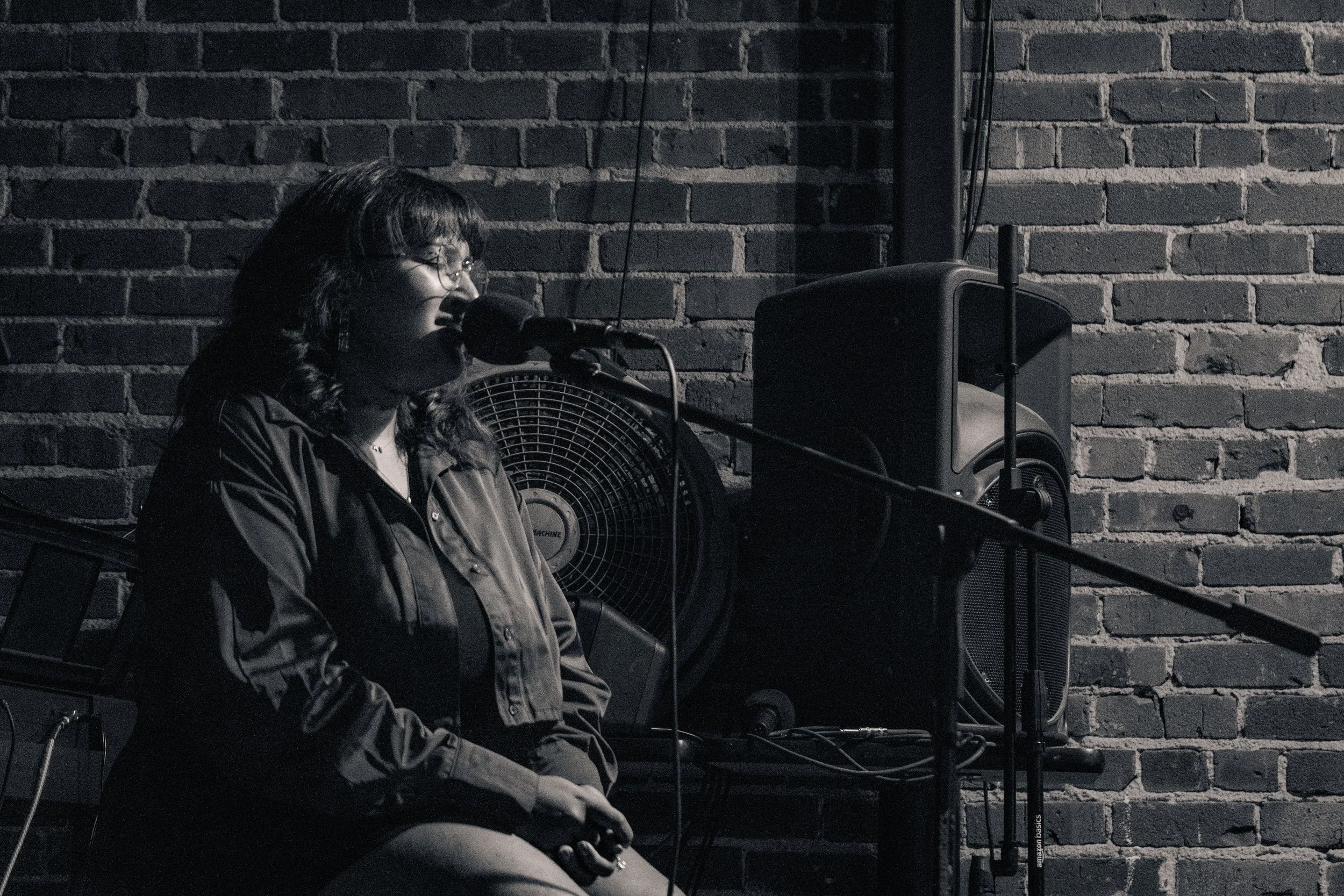 A woman with dark, wavy hair and glasses singing into a microphone on a stage with a brick wall background, accompanied by sound equipment and a fan.