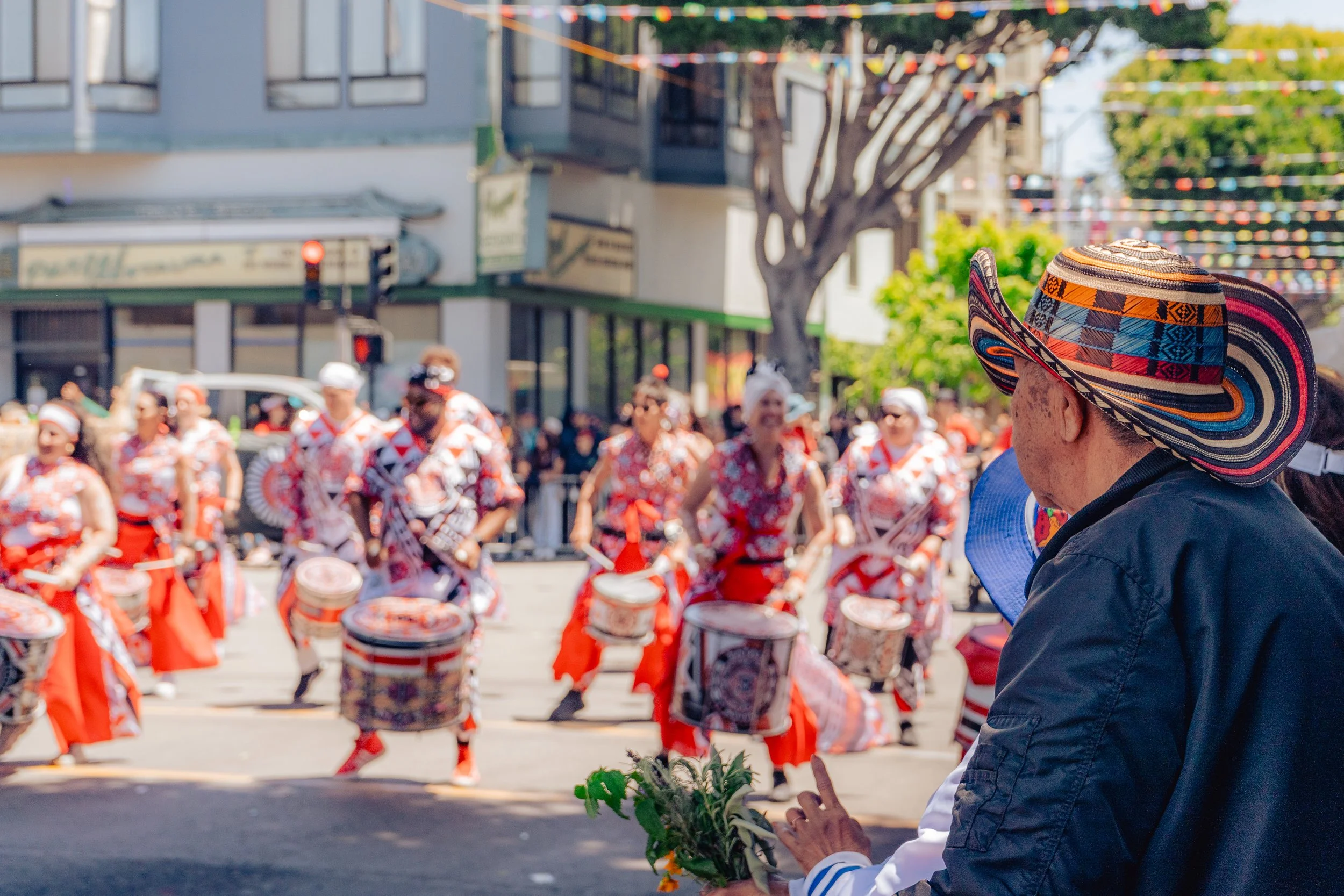 A person in a wide-brimmed, colorful hat watching a parade of performers in colorful costumes playing drums on a city street, with spectators and shops in the background.