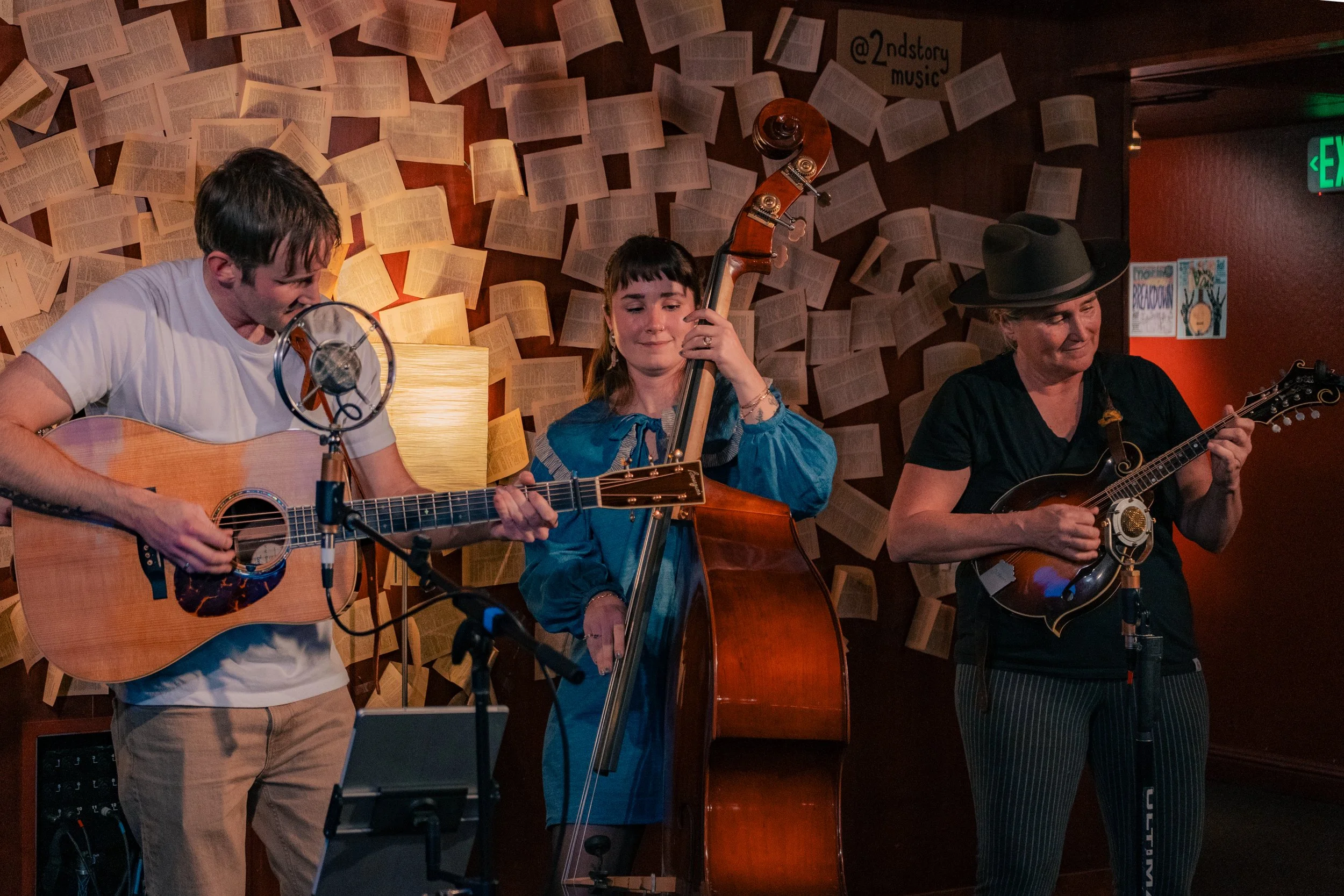 Three musicians performing on stage: a man playing an acoustic guitar and singing into a microphone, a woman playing a double bass, and another woman playing a mandolin, with a background of hanging pages or sheets of paper on the wall.