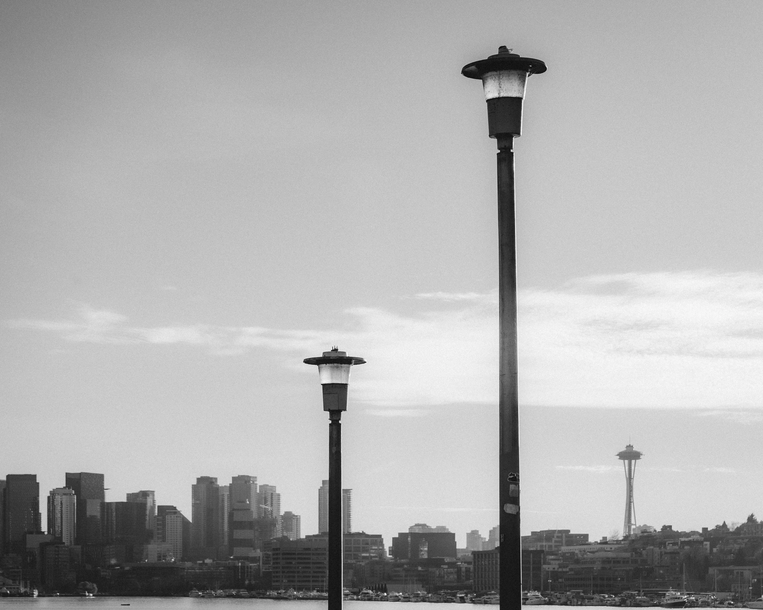 Black and white photo of city skyline with the Space Needle in the background, three tall street lamps in foreground, over water.