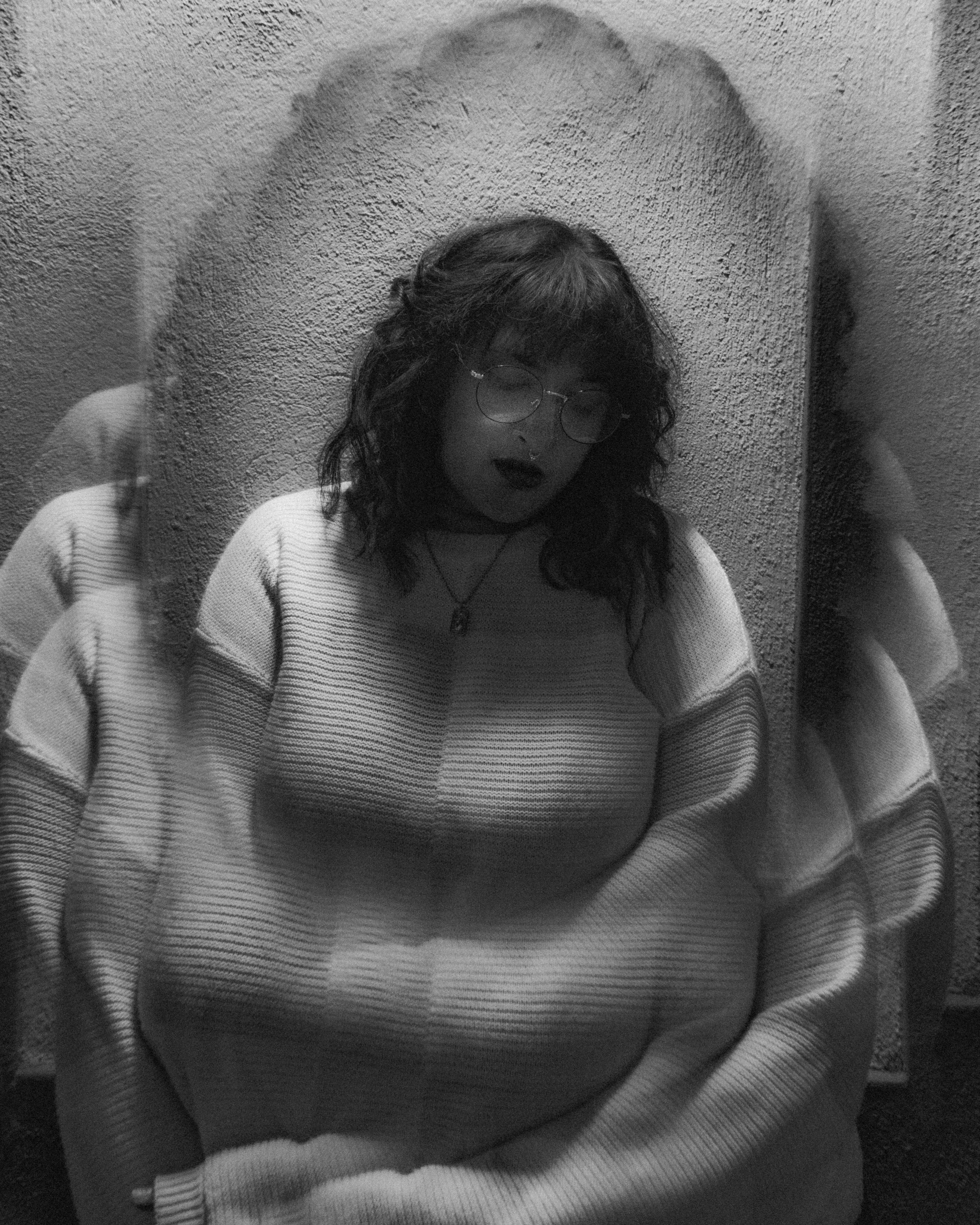 A woman with curly hair and glasses looking down, standing in front of a textured wall, reflected in a mirror.