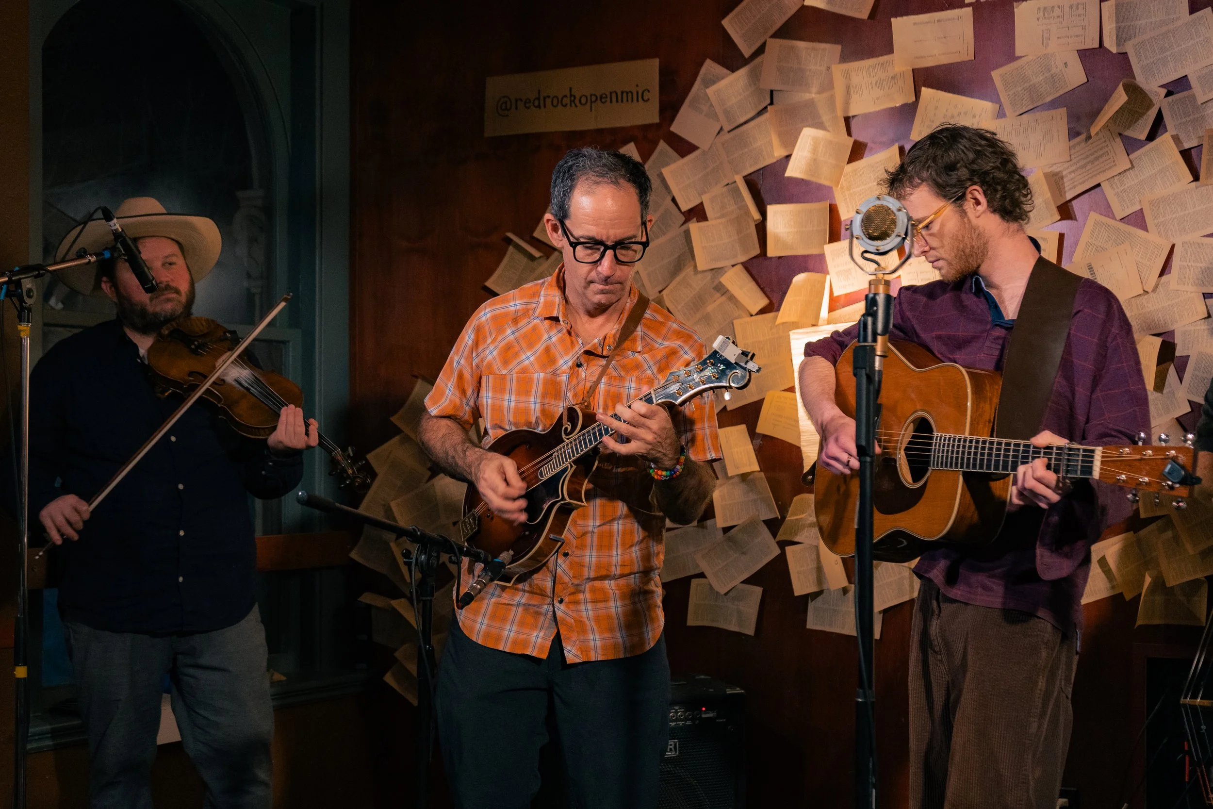 Three musicians performing indoors: a man on the left playing a violin, a man in the middle playing a mandolin, and a man on the right playing an acoustic guitar. The background features open books attached to the wall and a sign with the text '@redr