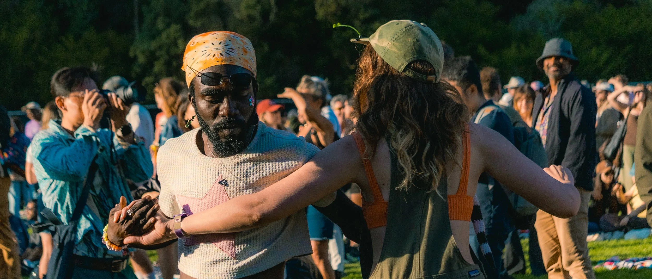 A group of diverse people outdoors, some dancing and others taking photos, at a social gathering or festival during daylight.