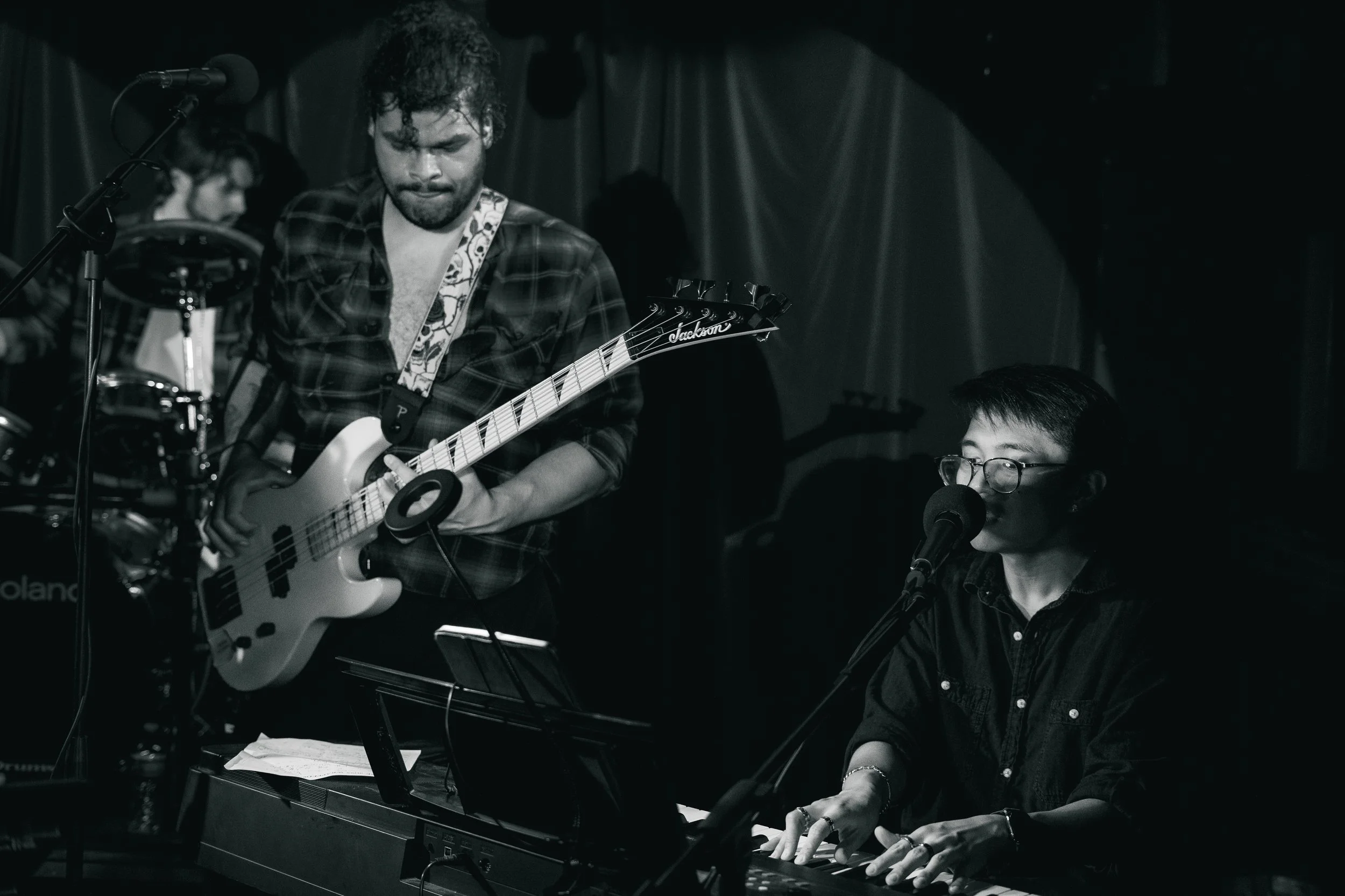 Two musicians performing on stage: one playing electric guitar, wearing a plaid shirt, and another playing keyboard and singing into a microphone, wearing glasses and a dark shirt, in a dimly lit setting.