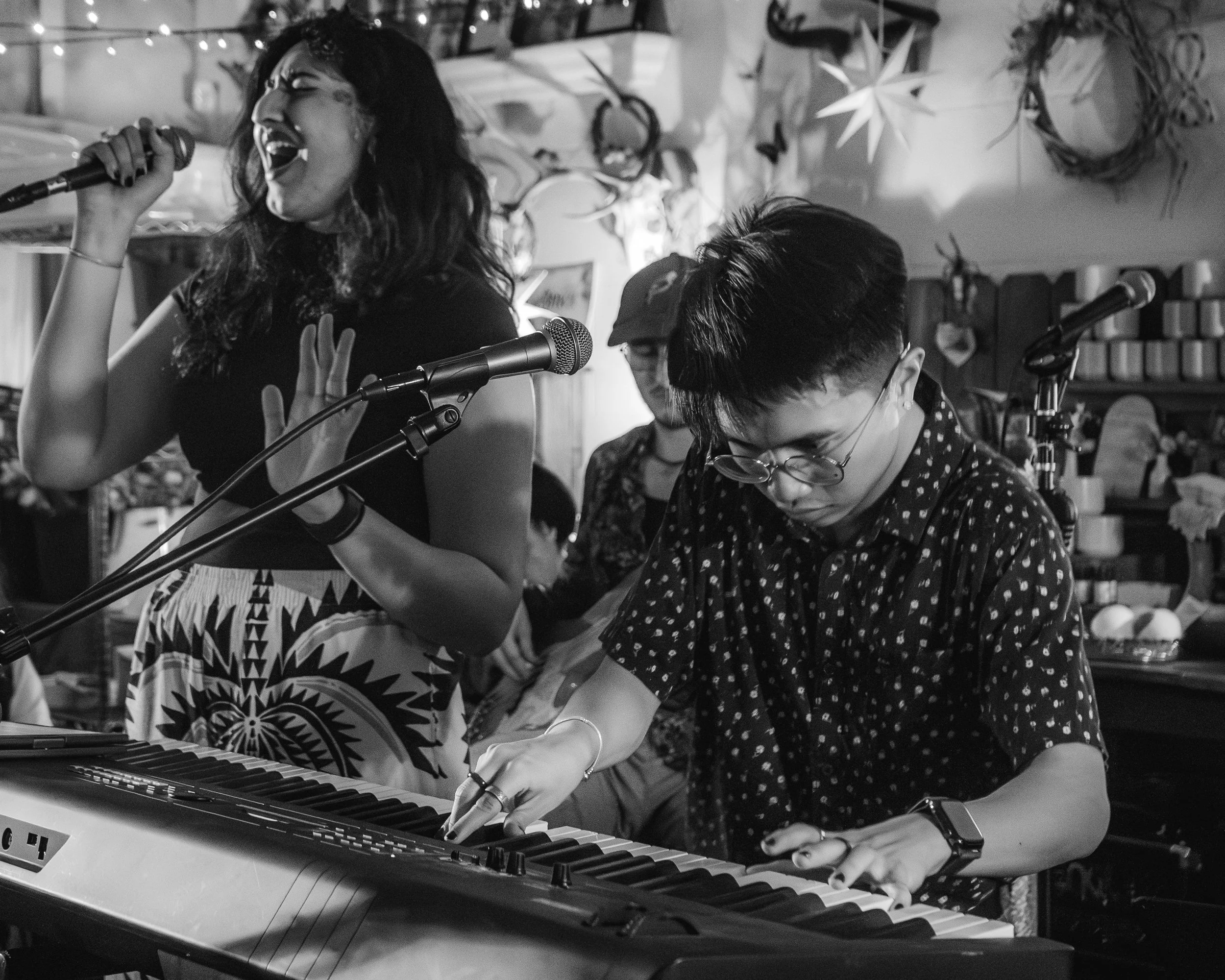 A woman passionately singing into a microphone while a man is playing a keyboard in a cozy, decorated room, with plants and hanging star decorations.