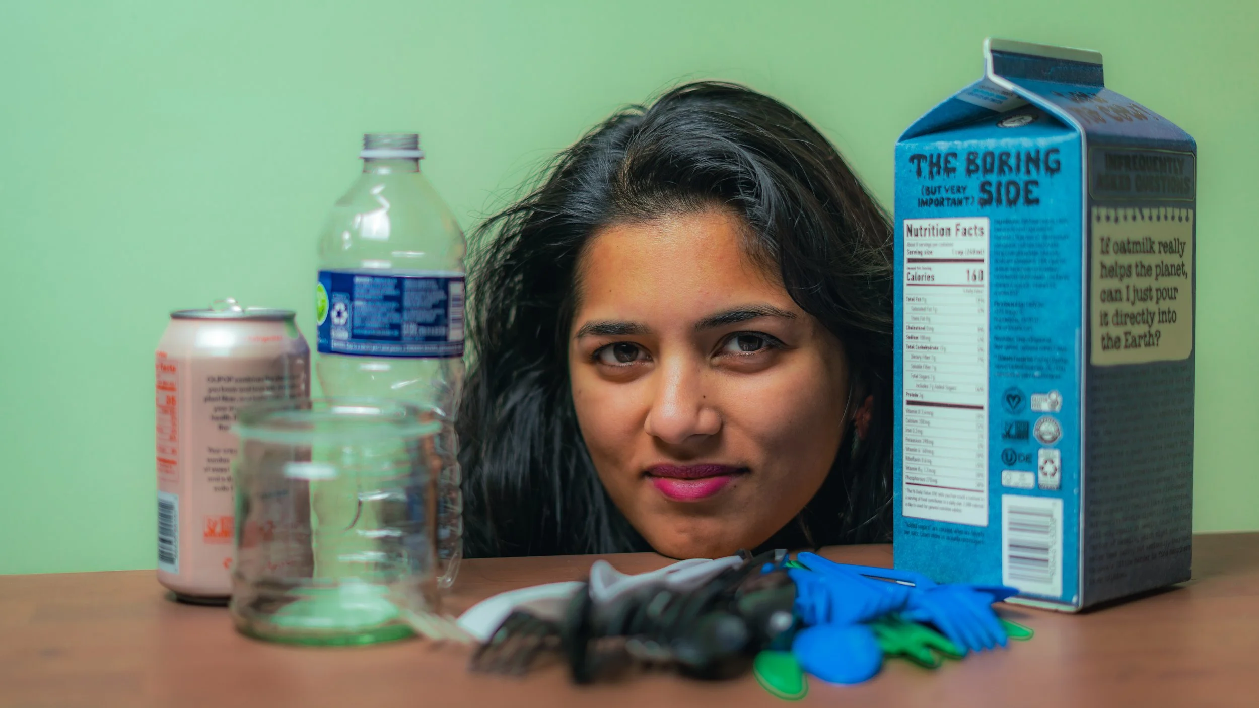 Woman with dark hair and pink lipstick leaning over a table surrounded by empty cans, bottles, a glass, gloves, and a cereal box with a humorous message.