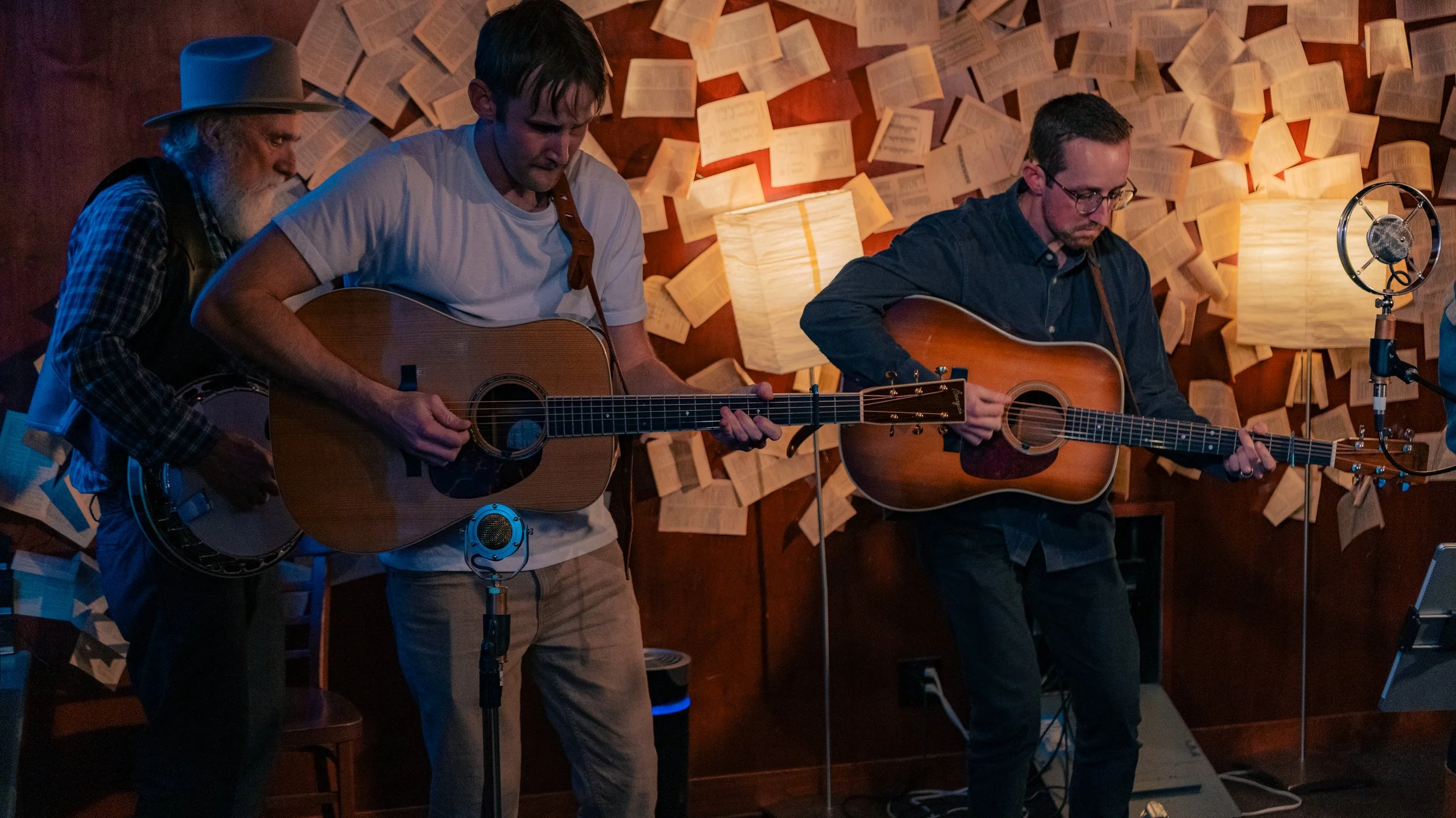 Three men playing guitars and a banjo in a cozy, dimly lit room with walls decorated with open books and warm lighting from lamps.