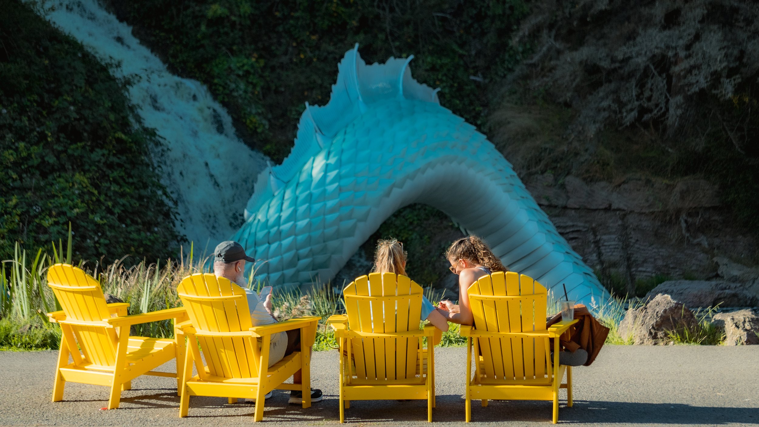 Three people sitting on yellow chairs in front of a large dragon-shaped sculpture, with a waterfall in the background.