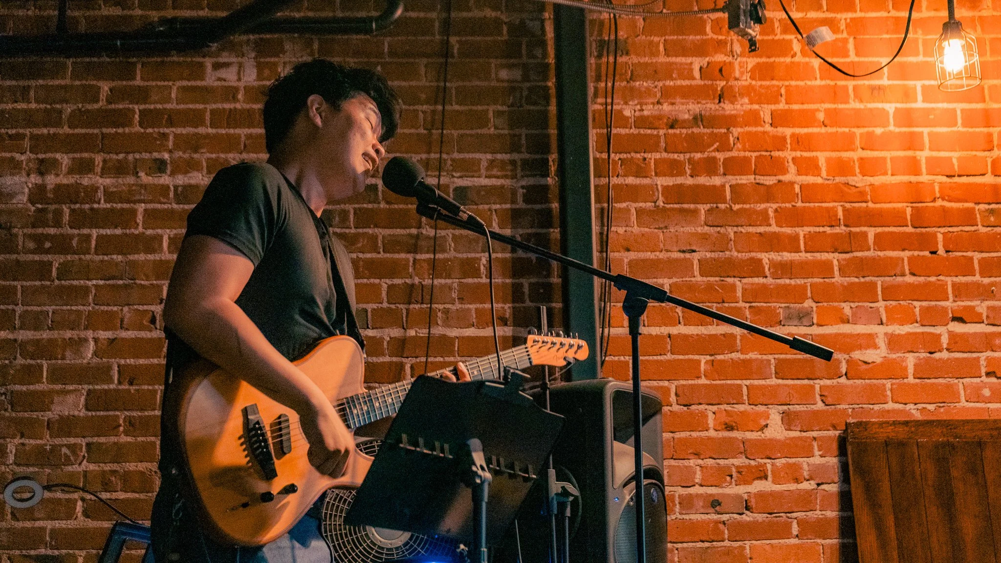 A male musician with dark hair playing an acoustic guitar in front of a red brick wall at a small performance venue, singing into a microphone with ambient lighting.