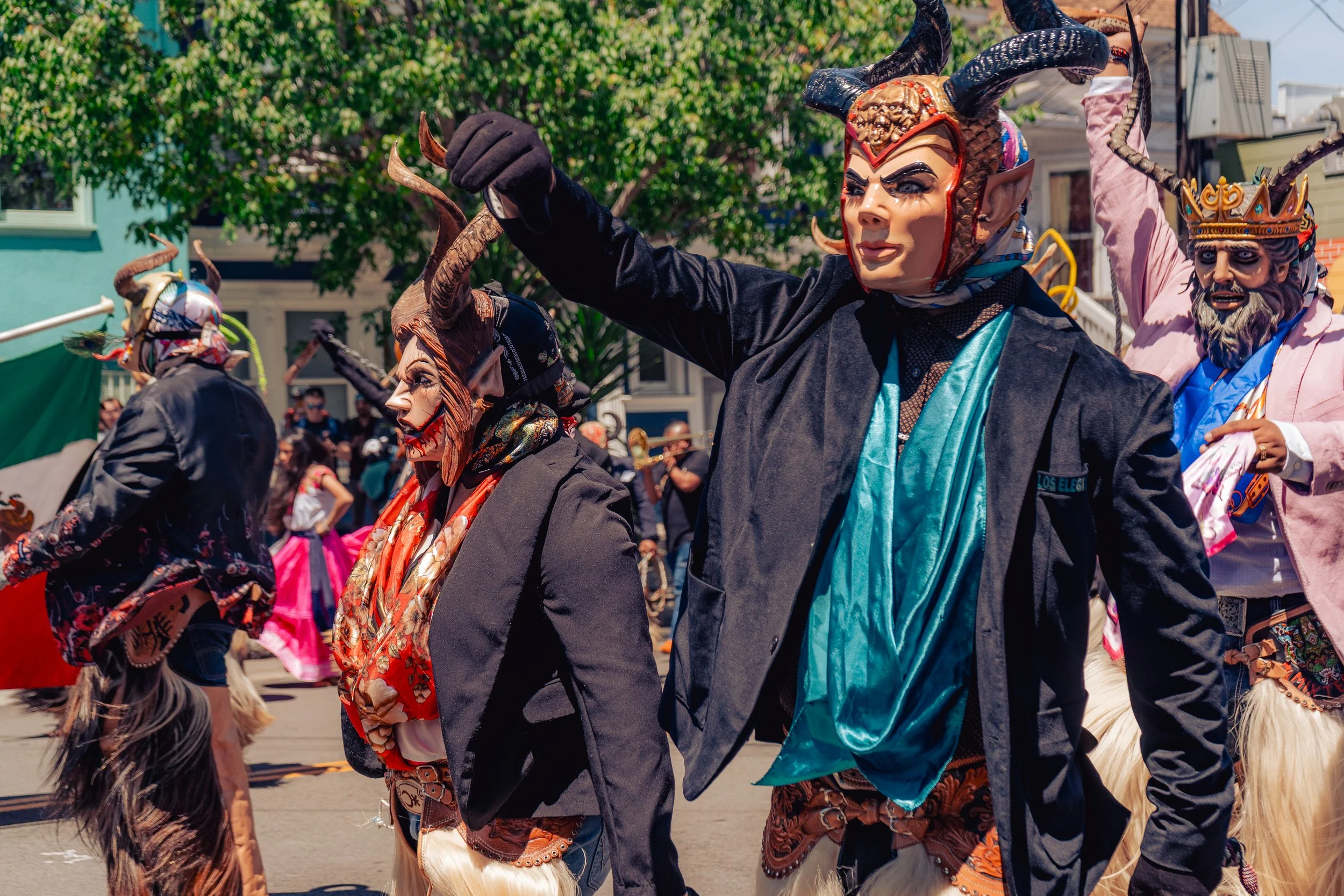 People wearing elaborate devil masks and costumes participating in a parade or festival on a sunny day, with trees and buildings in the background.