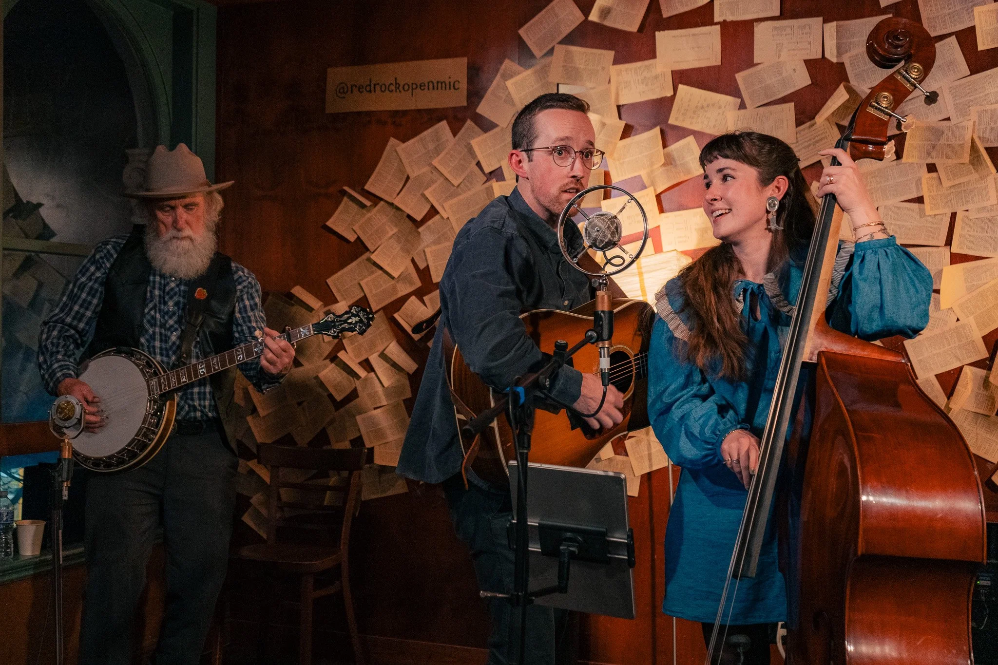 Three musicians performing in a cozy room with bookshelves. An older man with a white beard and hat playing a banjo, a man wearing glasses and a dark shirt playing an acoustic guitar, and a woman with long hair playing a double bass and smiling. The 