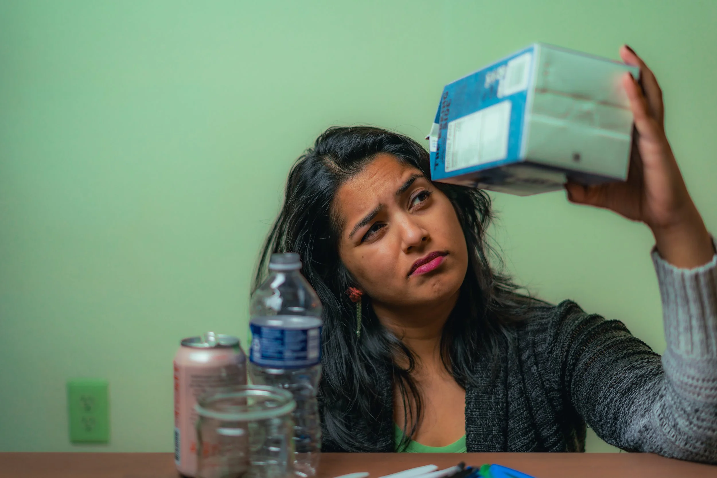 A woman sitting at a table holding a milk carton above her head with a curious expression, with bottles on the table in front of her.