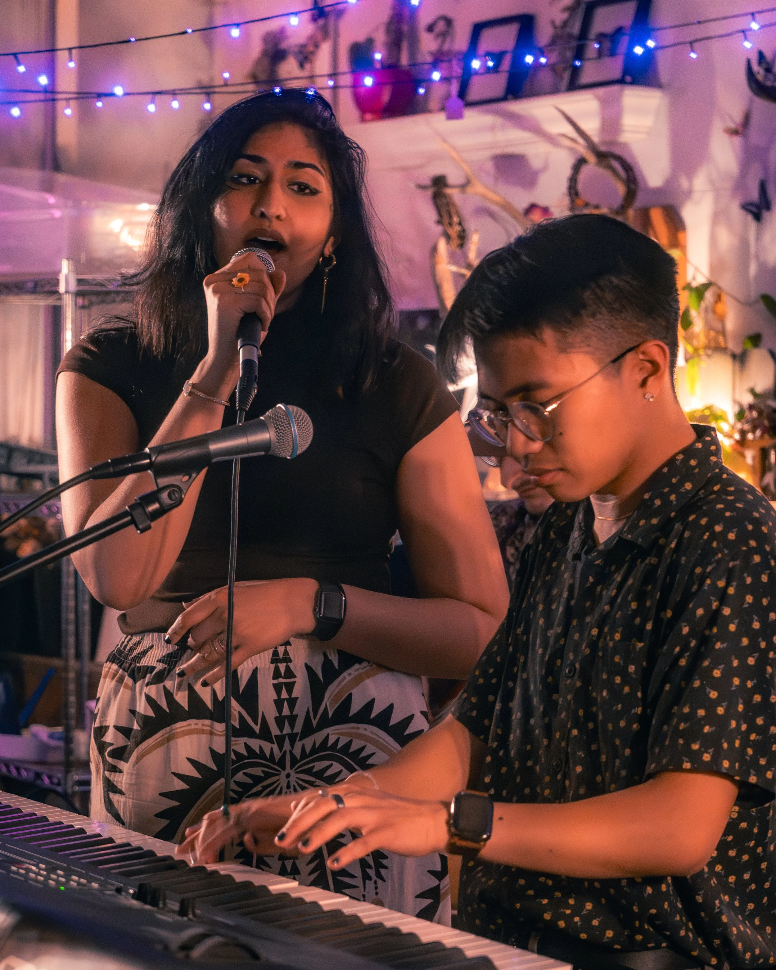 A woman and a person with glasses playing piano in a room decorated with string lights and plants, with the woman singing into a microphone.
