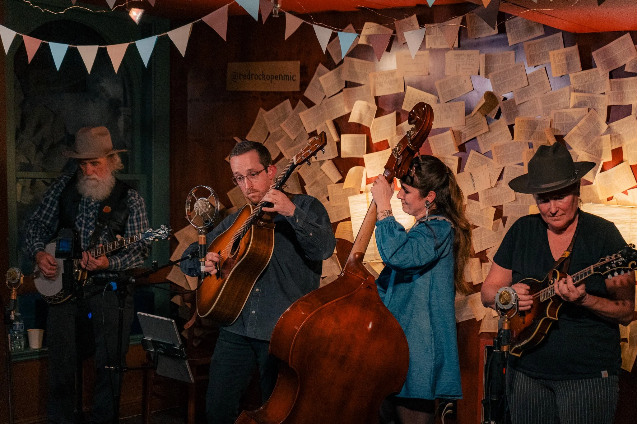 Four musicians playing guitars and a double bass on a stage with a wall decorated with open books and string lights.