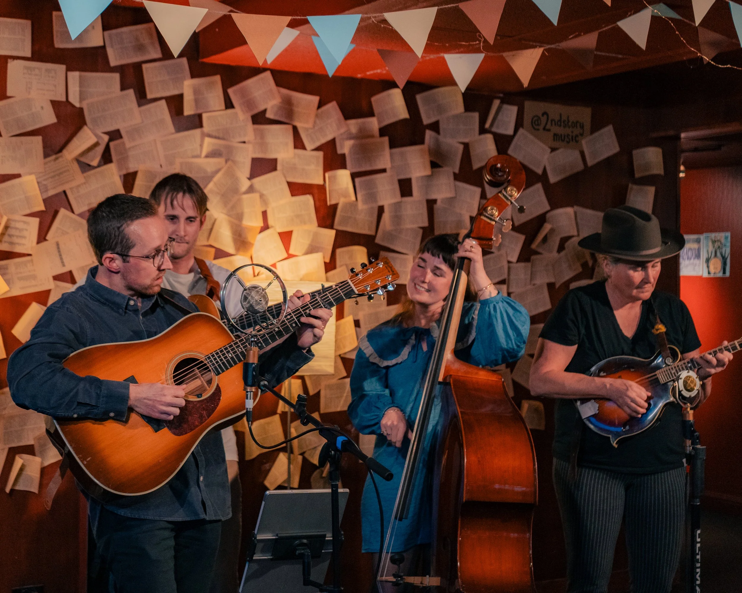 Four musicians playing instruments on stage, with a wall of open books behind them and paper bunting overhead.