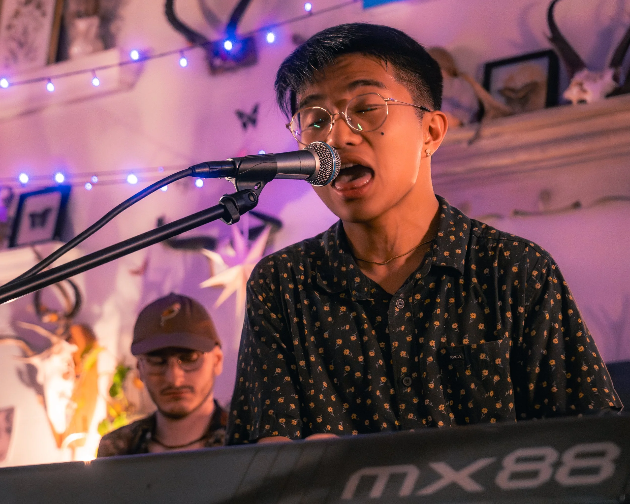 A person singing into a microphone and playing an electronic keyboard, with a person in the background, decorated with string lights, framed pictures, and mounted animal horns.
