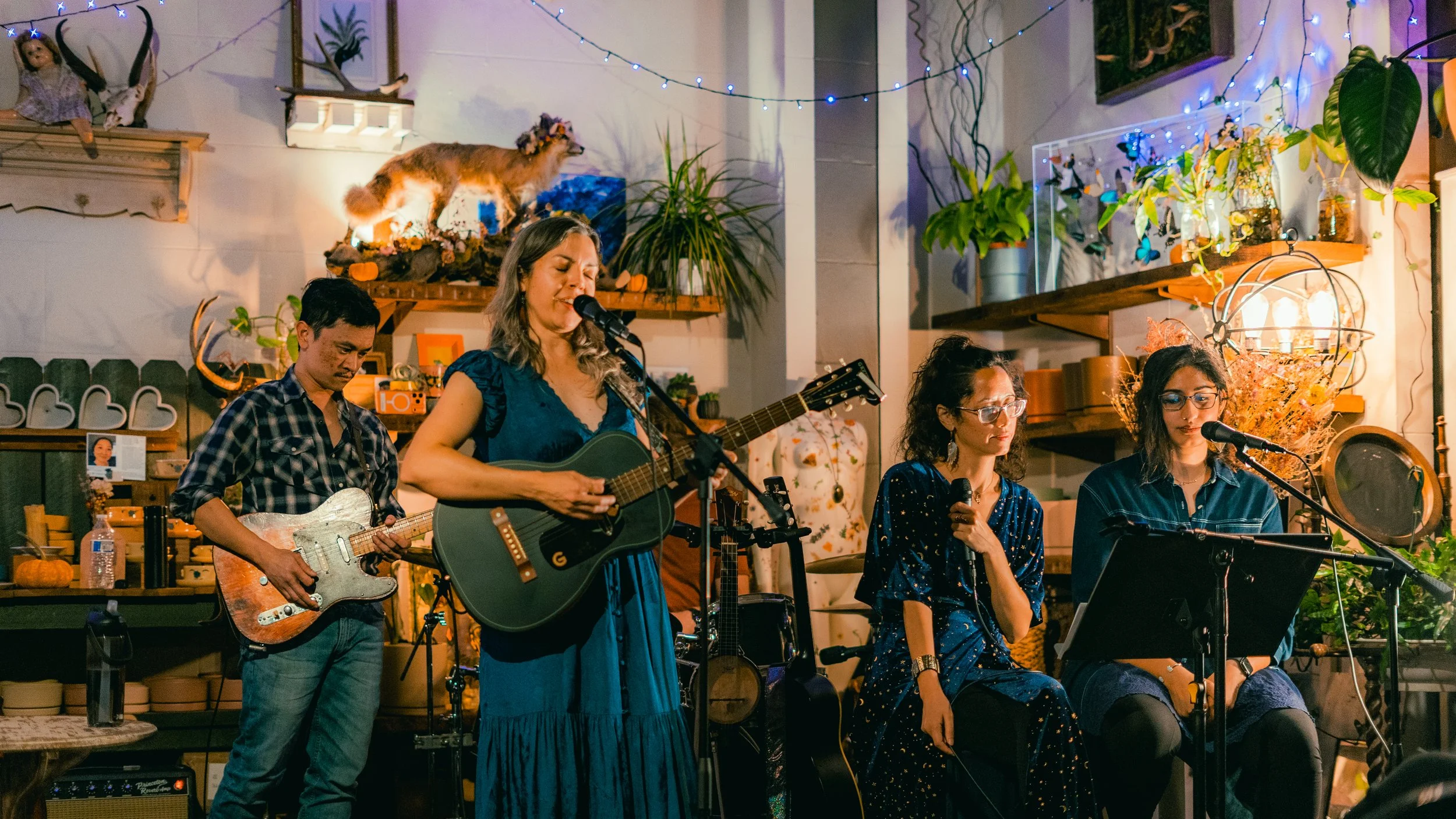 Four musicians performing in a cozy, decorated space: a woman playing guitar and singing, a man playing electric guitar, and two women sitting with microphones, one singing and one reading sheet music. The background features plants, string lights, a
