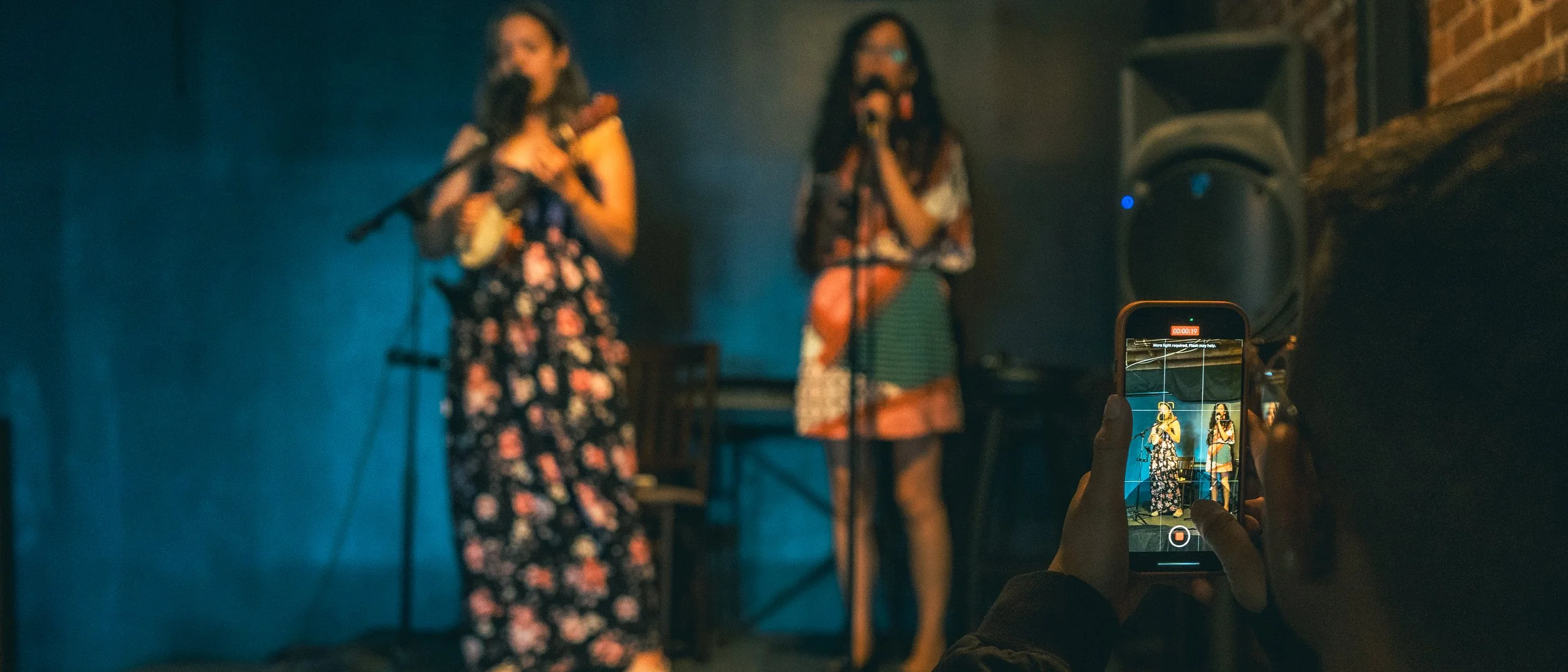 Two women are performing on stage, one playing a flute and the other singing into a microphone, with a person recording their performance on a smartphone in the foreground.