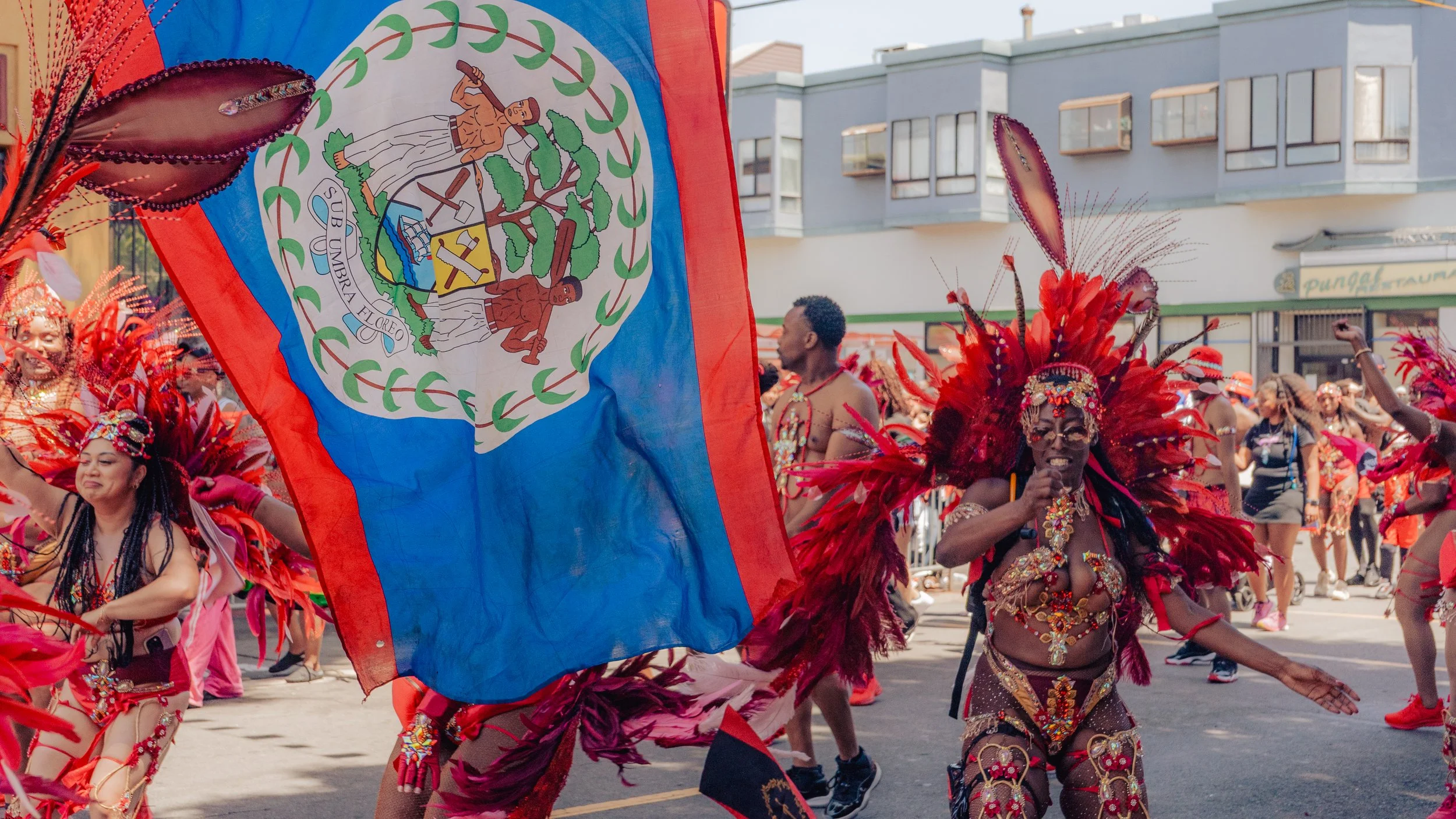 People in colorful costumes dancing during a parade, with a woman in elaborate red feathered headdress and accessories prominently featured. A large Belize flag is being waved among the crowd.