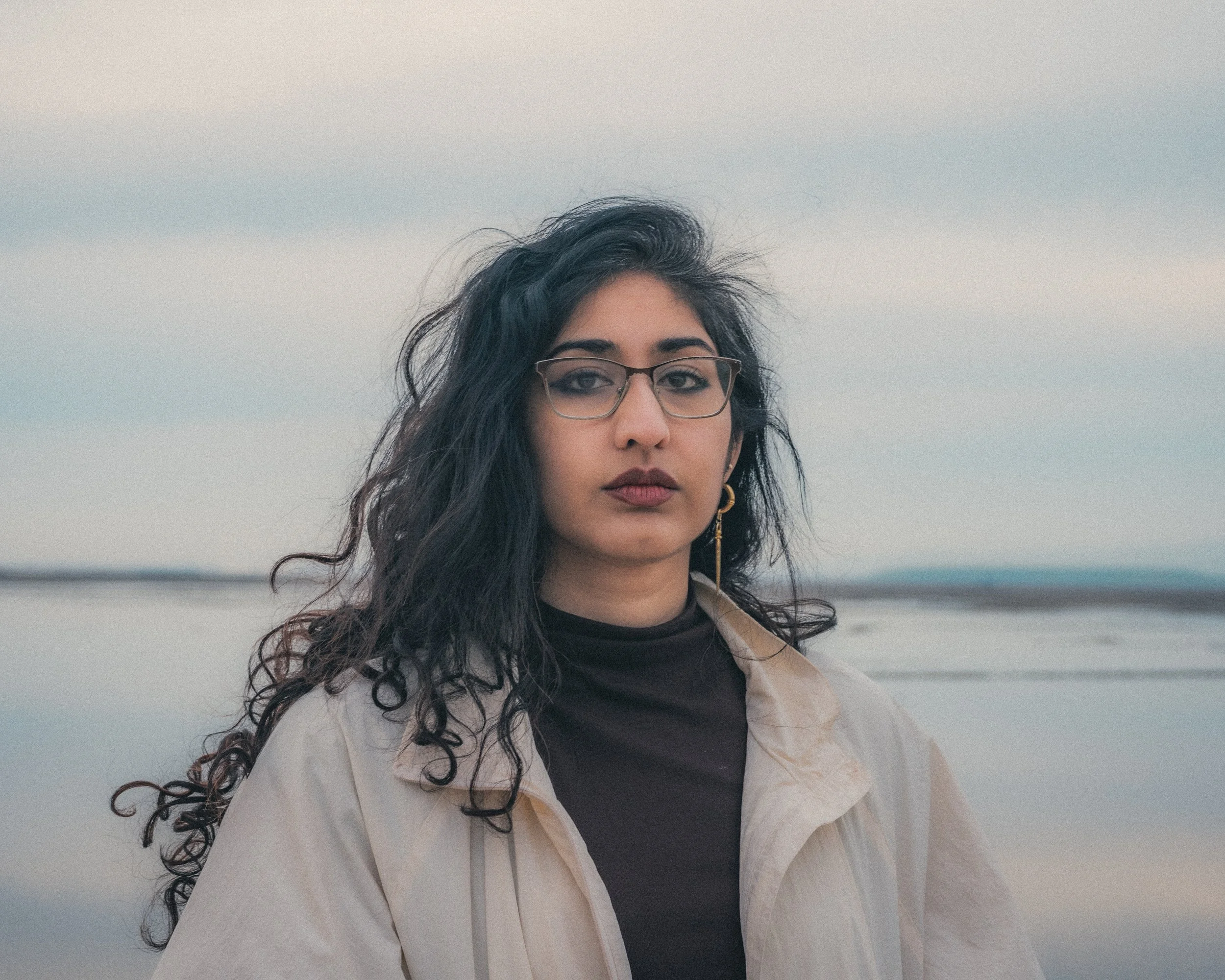 A woman with long curly black hair, glasses, and gold earrings standing outdoors during daytime with a body of water and cloudy sky in the background.